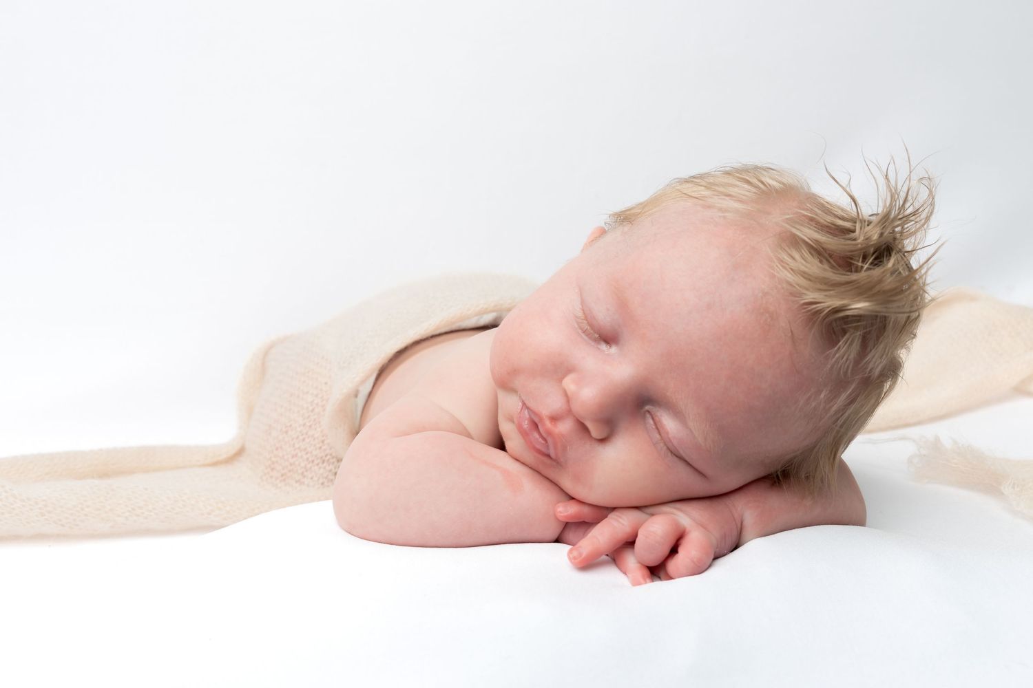 Peaceful newborn baby sleeping on white background, resting chin on hands, wrapped in a soft beige blanket.