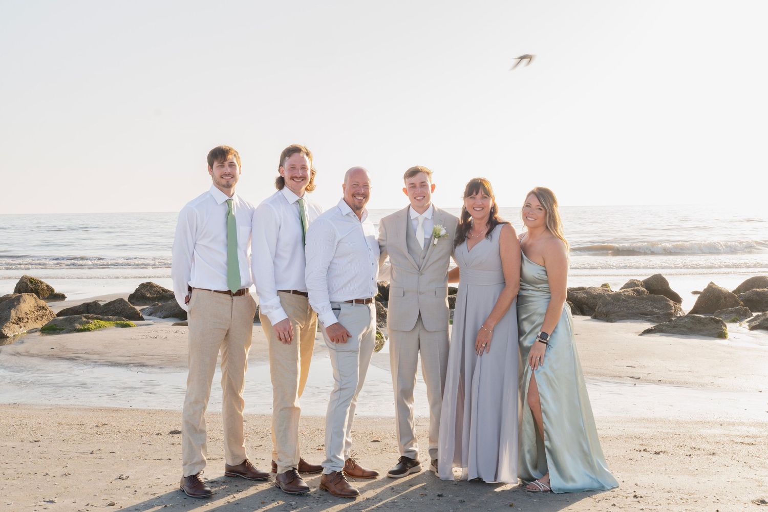 Wedding party posing on a sunny beach, groomsmen in khaki and bridesmaids in sage green dresses.