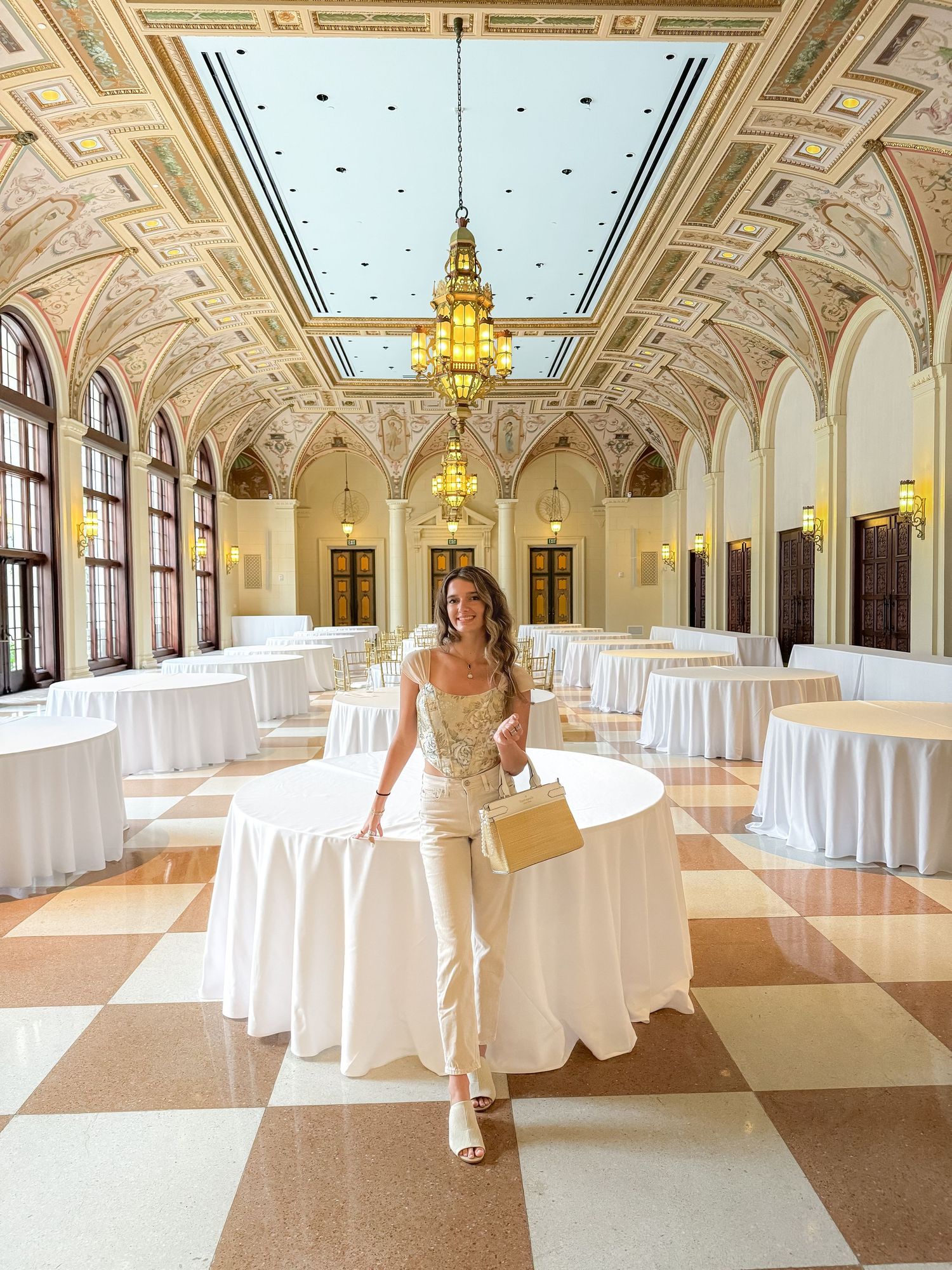 A woman in white walks through an ornate ballroom with arched ceilings and chandeliers.