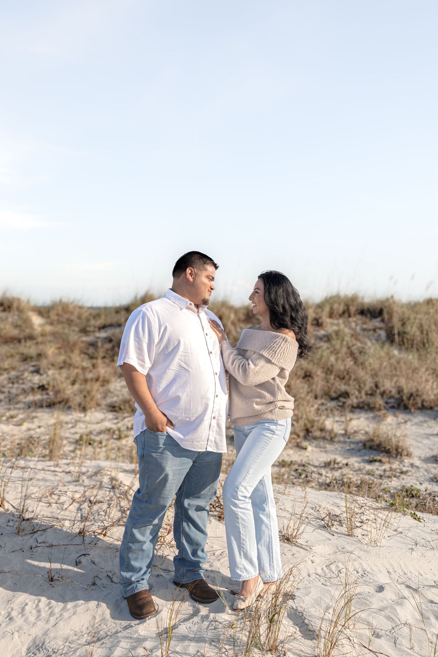 Couple standing together on sandy beach dunes, woman in beige sweater and jeans, man in white shirt, gazing at each other.