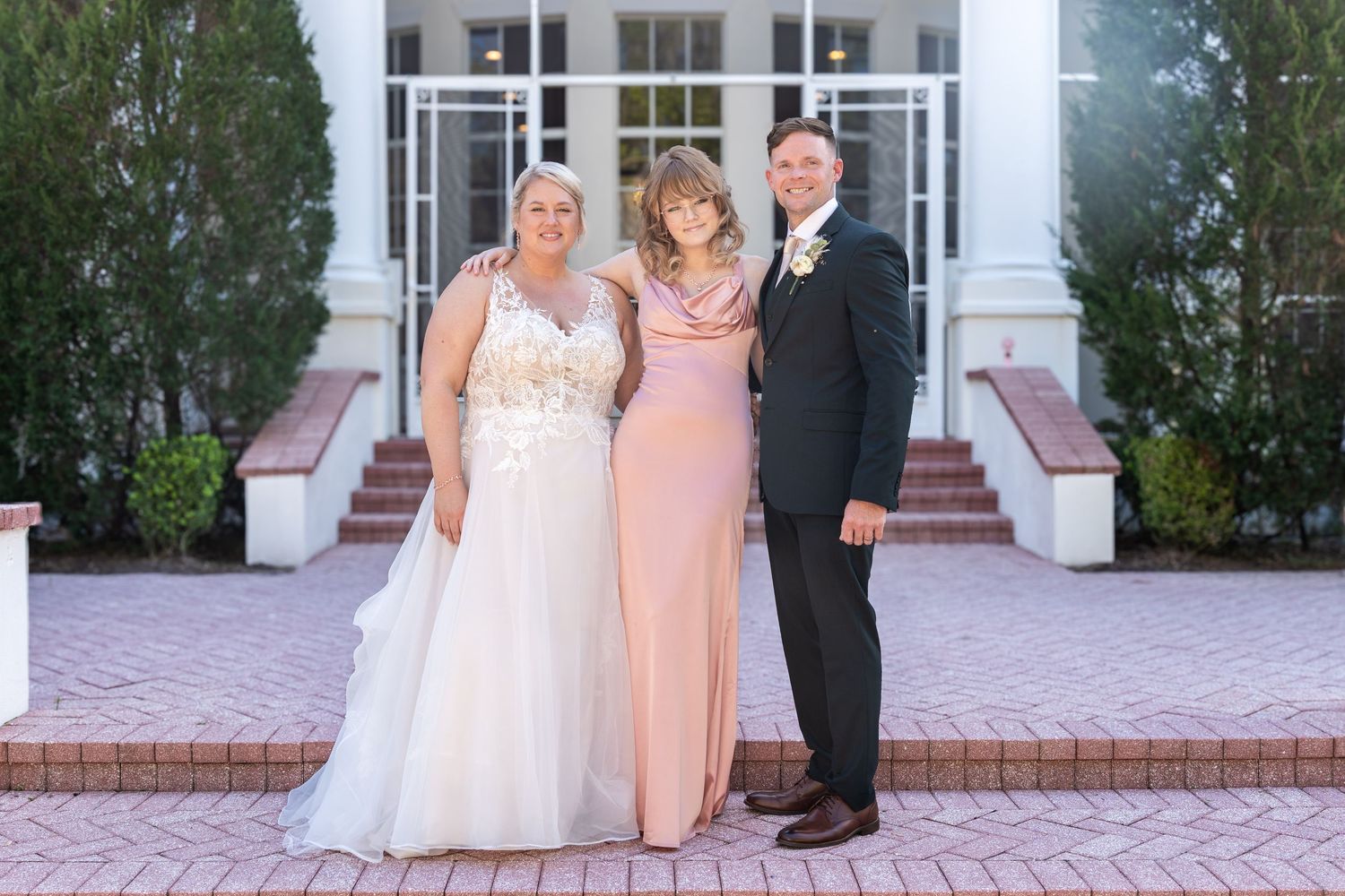 Bride in white gown poses with woman in pink dress and groom in dark suit outside elegant wedding venue.