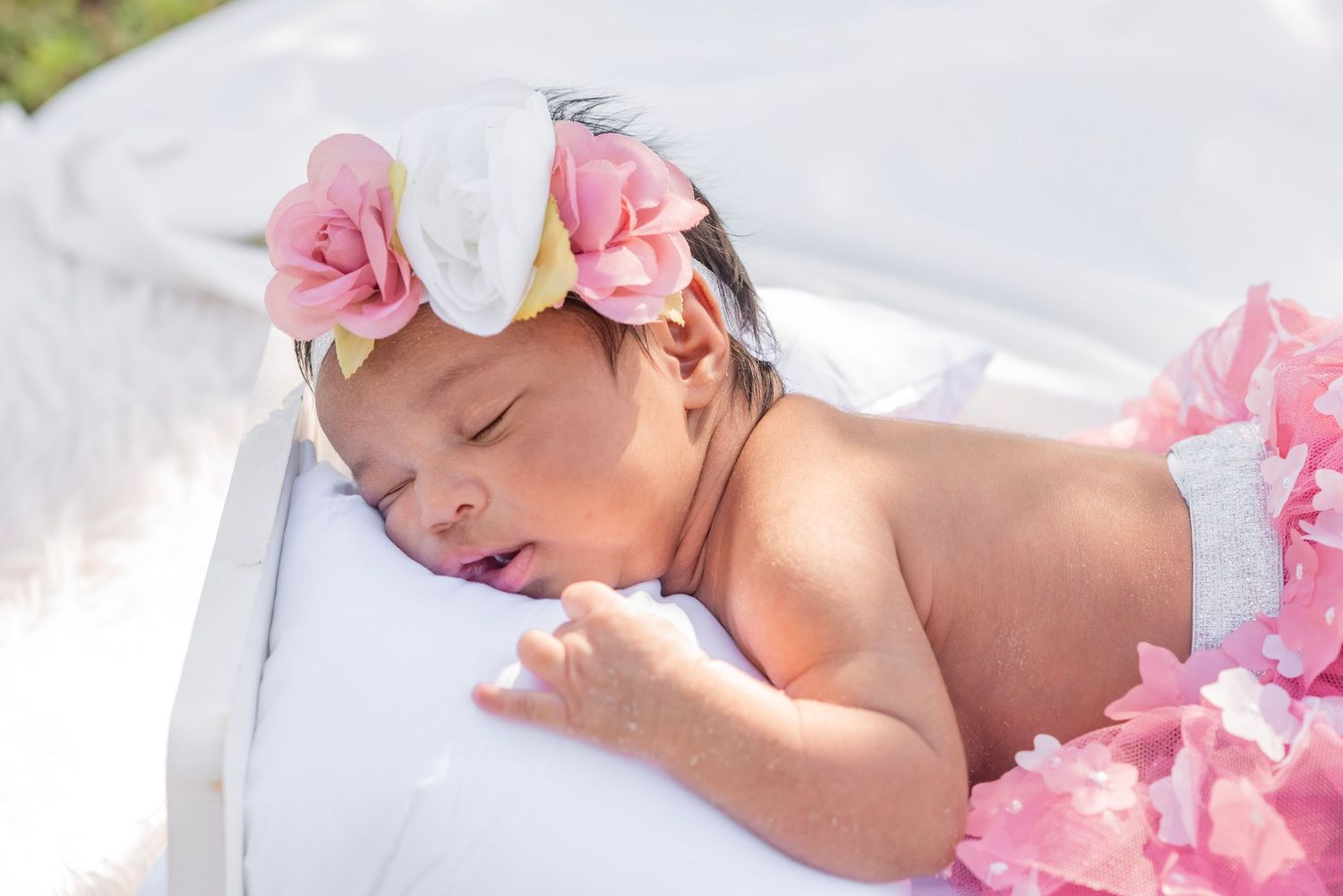 Sleeping newborn baby girl wearing a pink floral headband and pink ruffled outfit, resting peacefully on a white pillow.