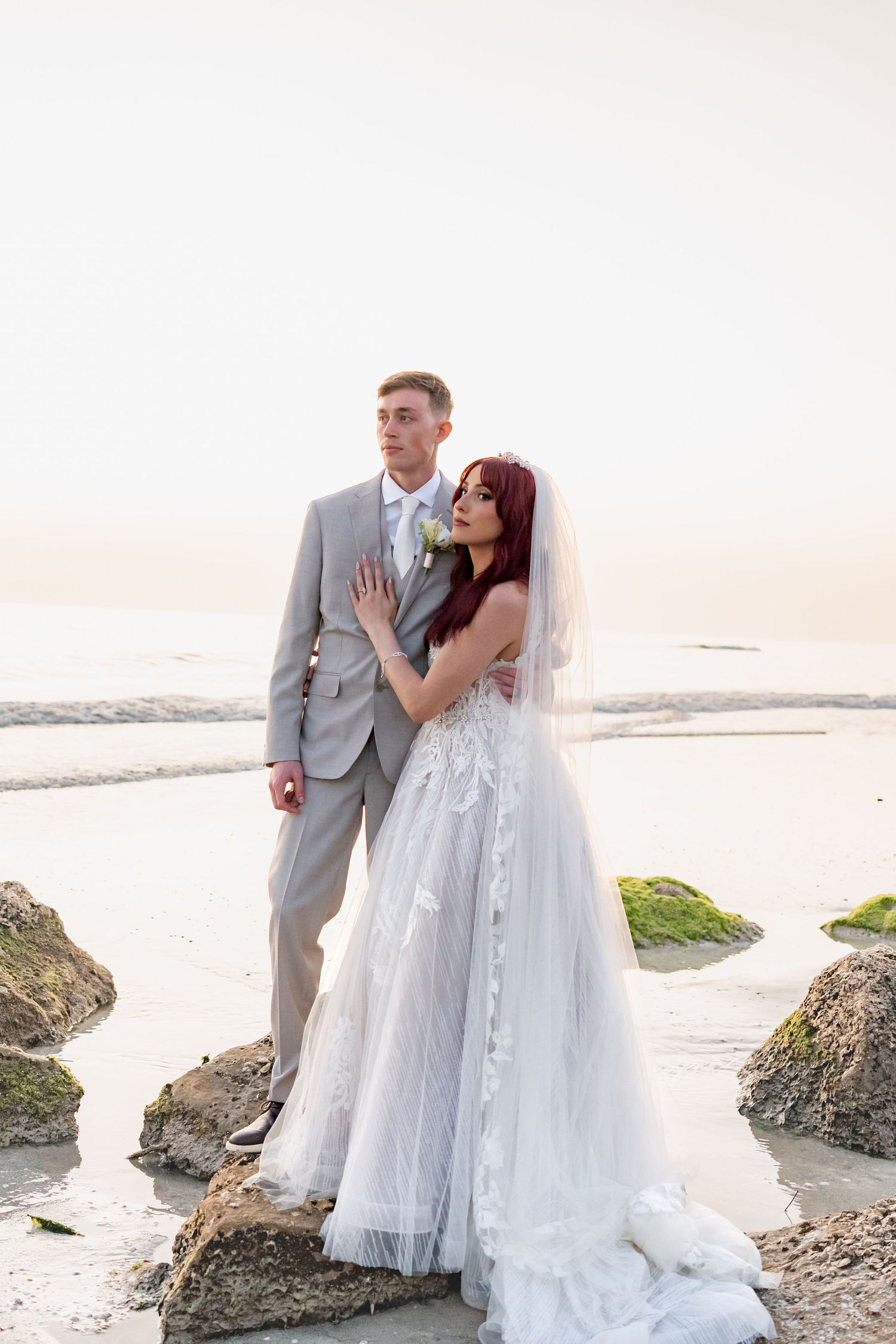 Bride and groom posing on rocky beach at sunset, bride in lace gown with veil, groom in gray suit.