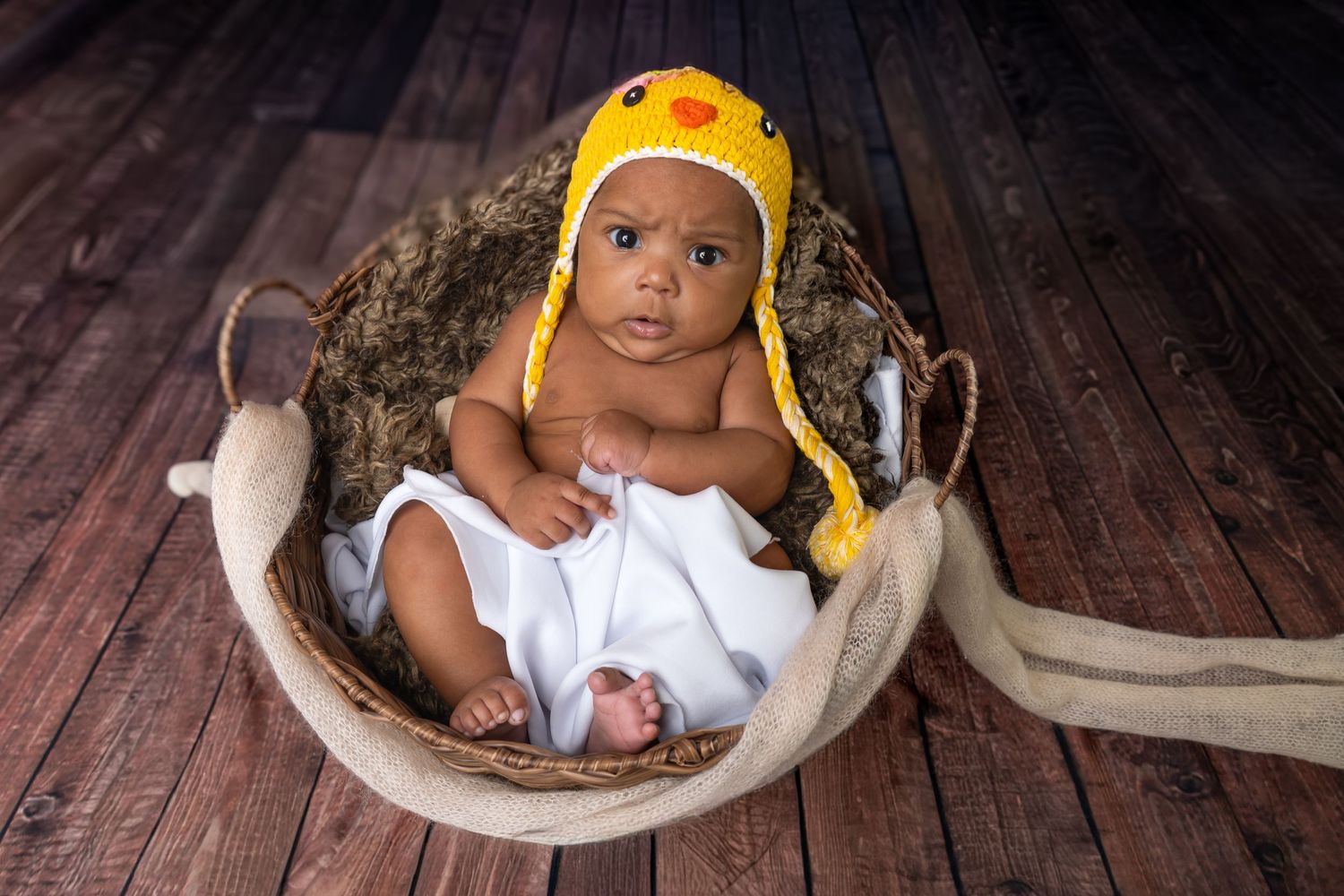 Adorable newborn baby wearing yellow crochet chick hat sitting in rustic basket with white blanket on wood floor.