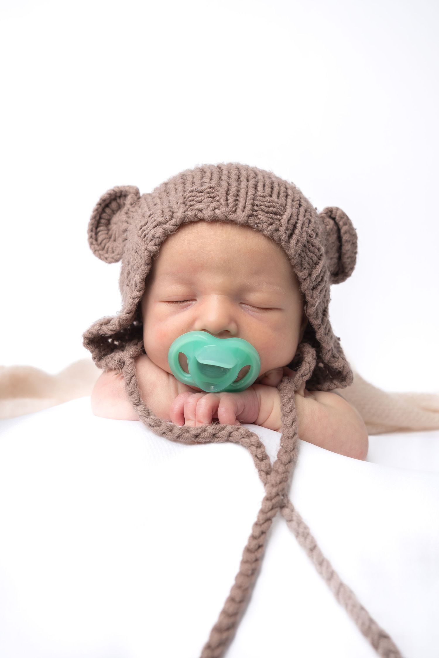 Sleeping newborn baby wearing a brown knitted bear hat with ears and a teal pacifier on a white background.