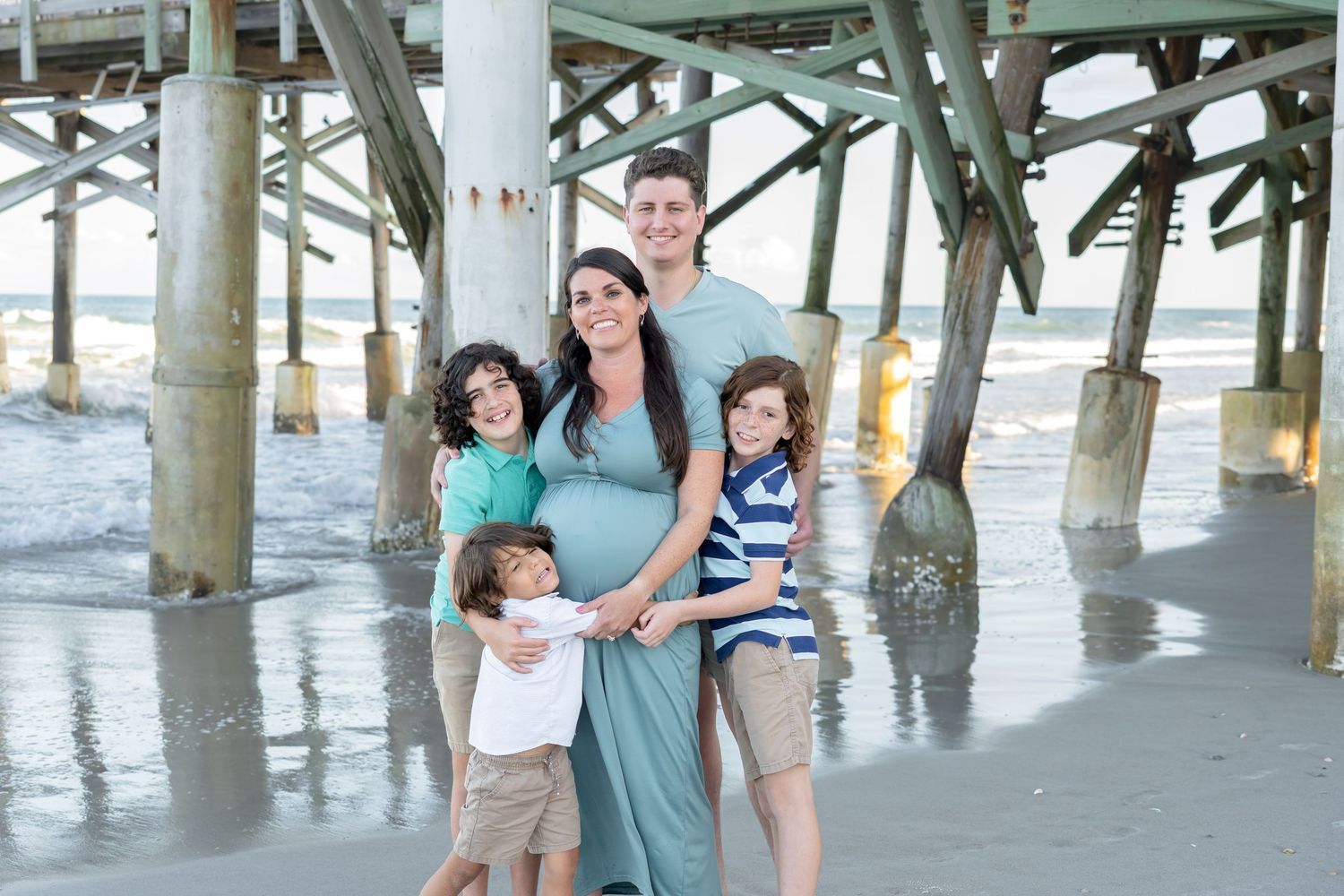 Family of five poses under a beach pier, with pregnant mother and smiling children on a sunny day.