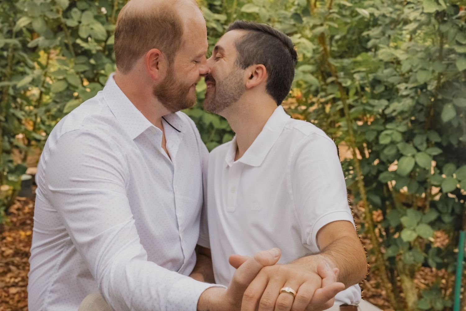 Two men in white shirts share a romantic moment outdoors, one wearing a wedding ring, surrounded by lush green foliage.