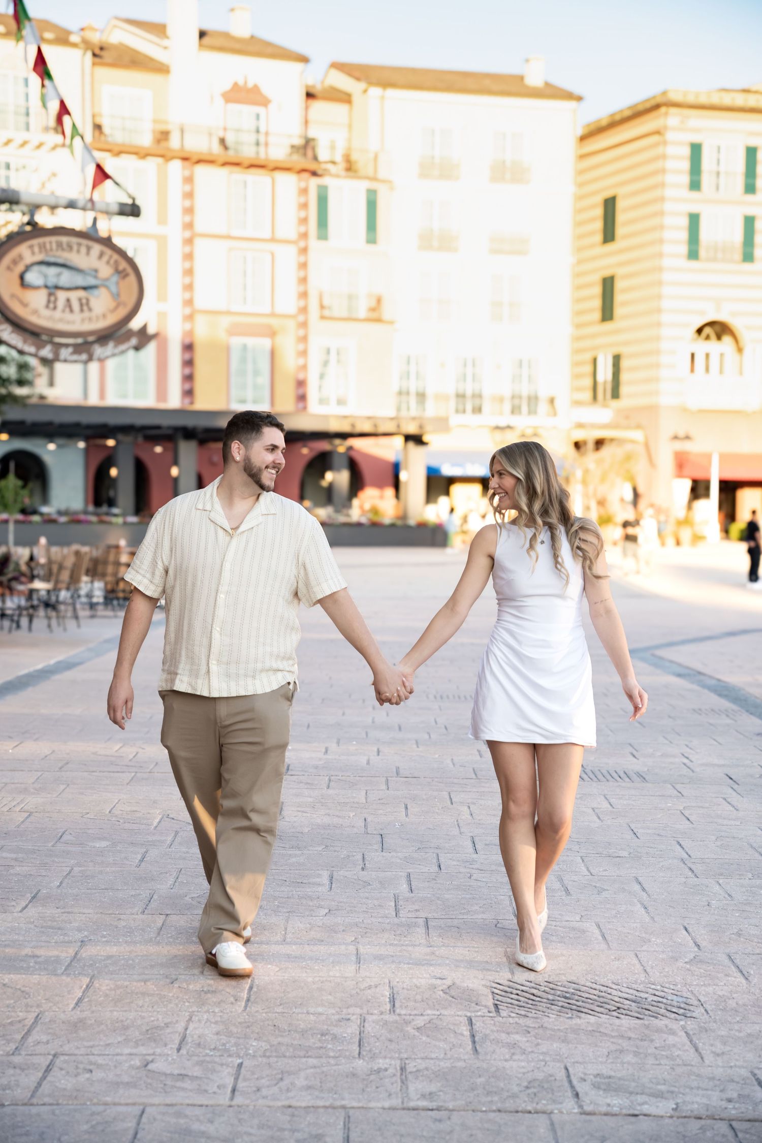 A smiling couple holds hands while walking through a sunny European-style plaza, man in beige shirt and woman in white dress.