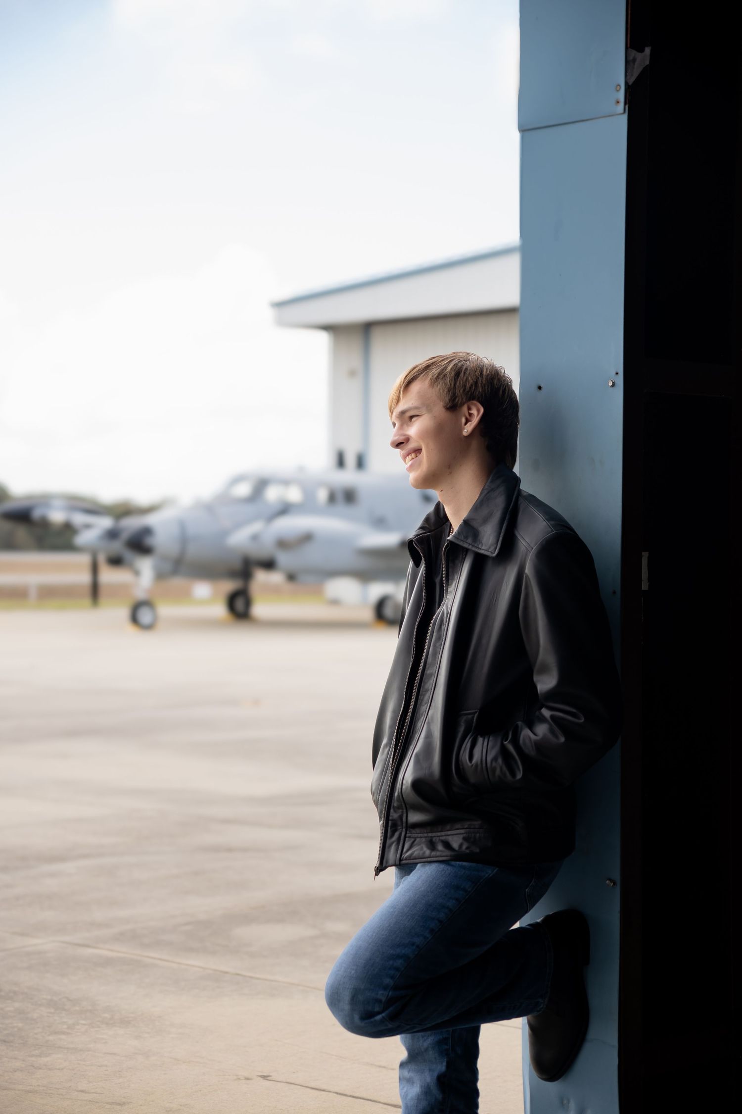 Young man in black leather jacket smiling outdoors near aircraft hangar with military jet in background.