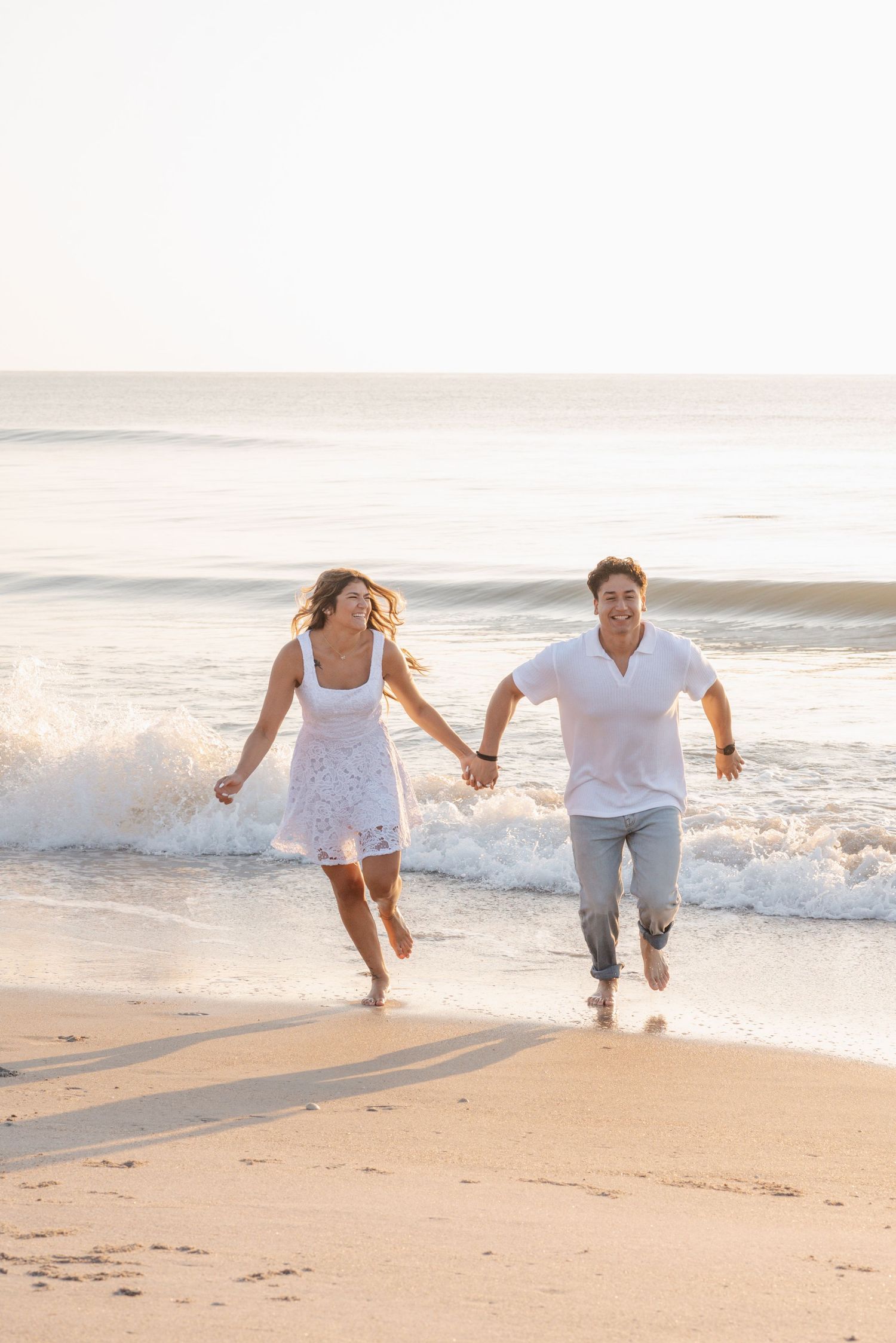 Joyful couple running hand-in-hand along a sunny beach shoreline with ocean waves behind them.
