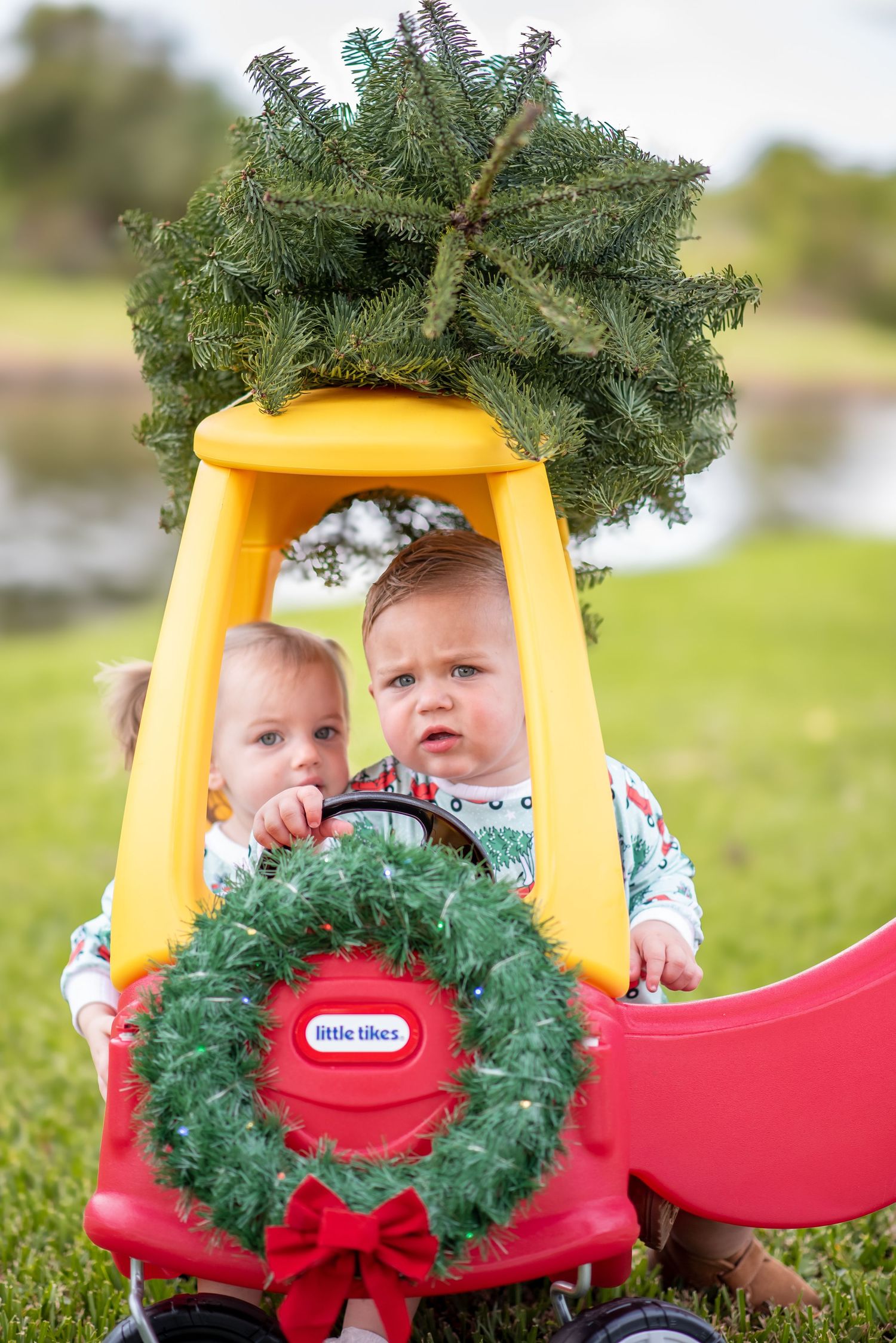 Two babies in a Little Tikes car decorated with a Christmas wreath and tree on top, outdoors on green grass.