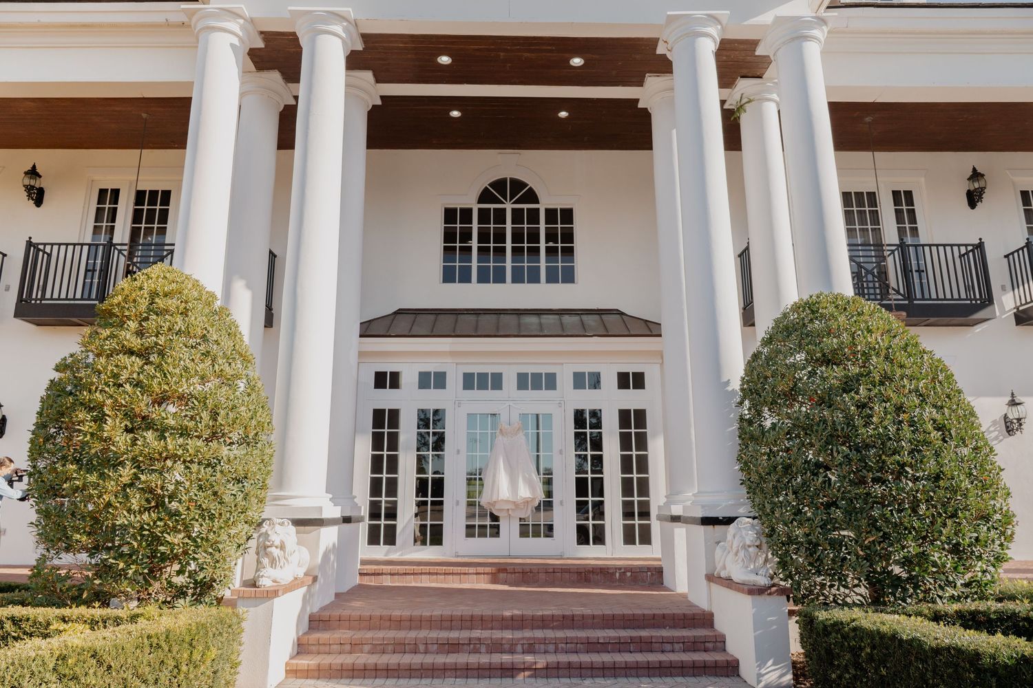 Grand estate venue entrance with tall white columns, manicured hedges, and French doors adorned with a wedding dress.