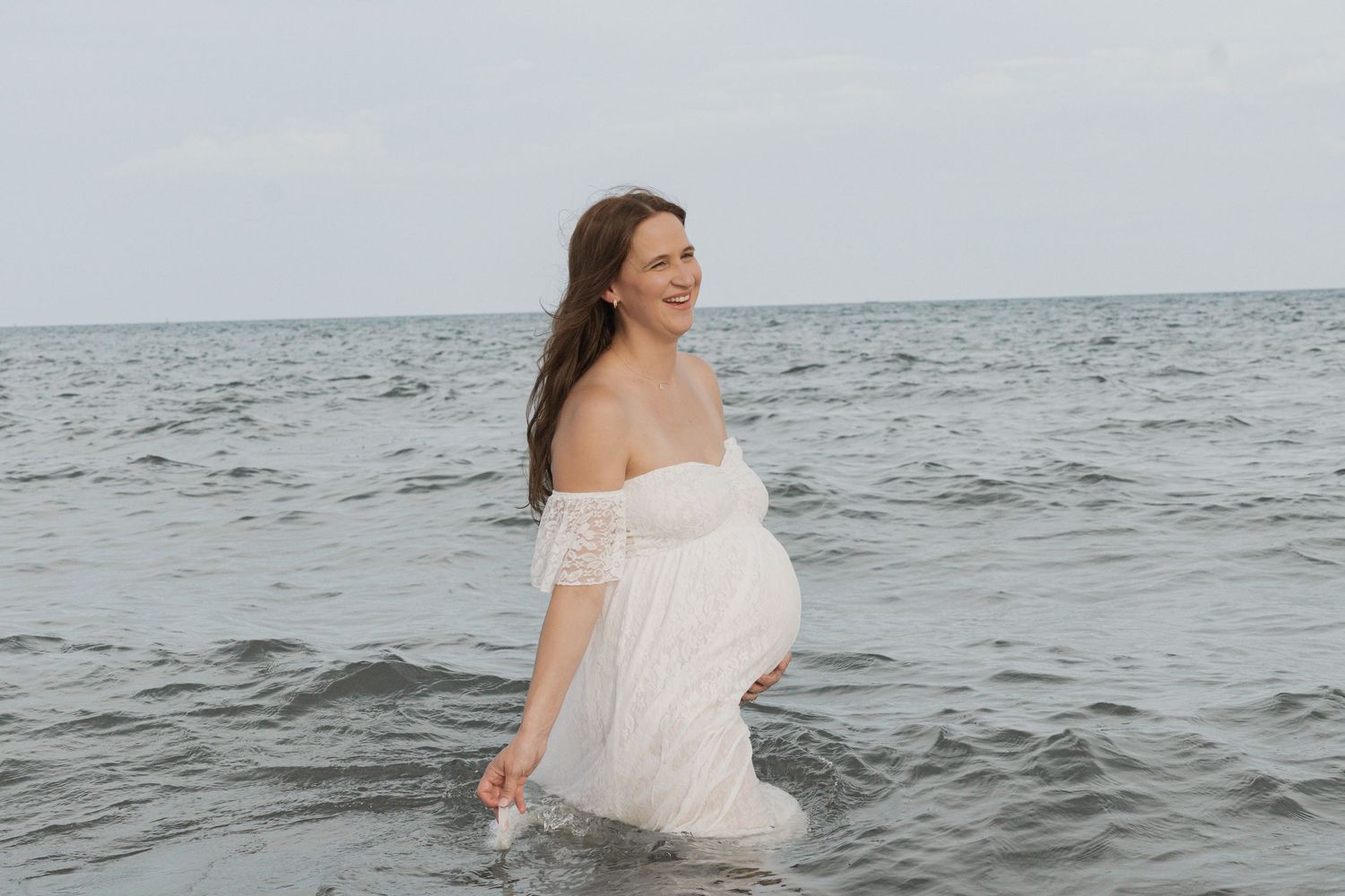 Pregnant woman in white off-shoulder lace dress wading in ocean waves, smiling joyfully during maternity photoshoot.