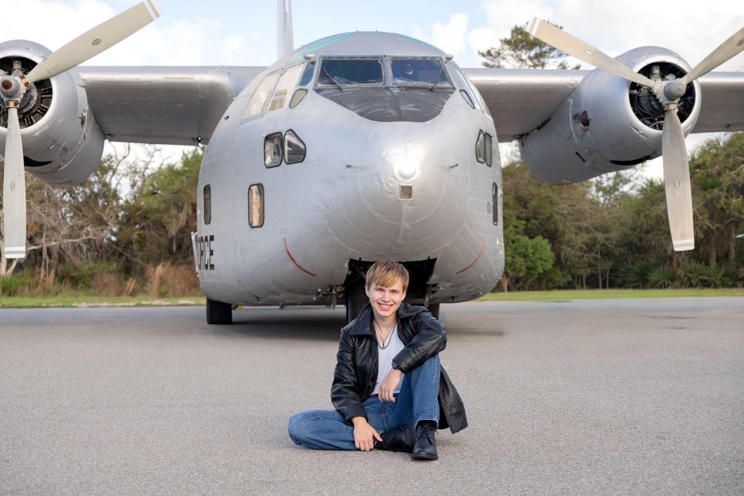 Young person in dark jacket sits cross-legged on tarmac in front of a large military propeller aircraft.