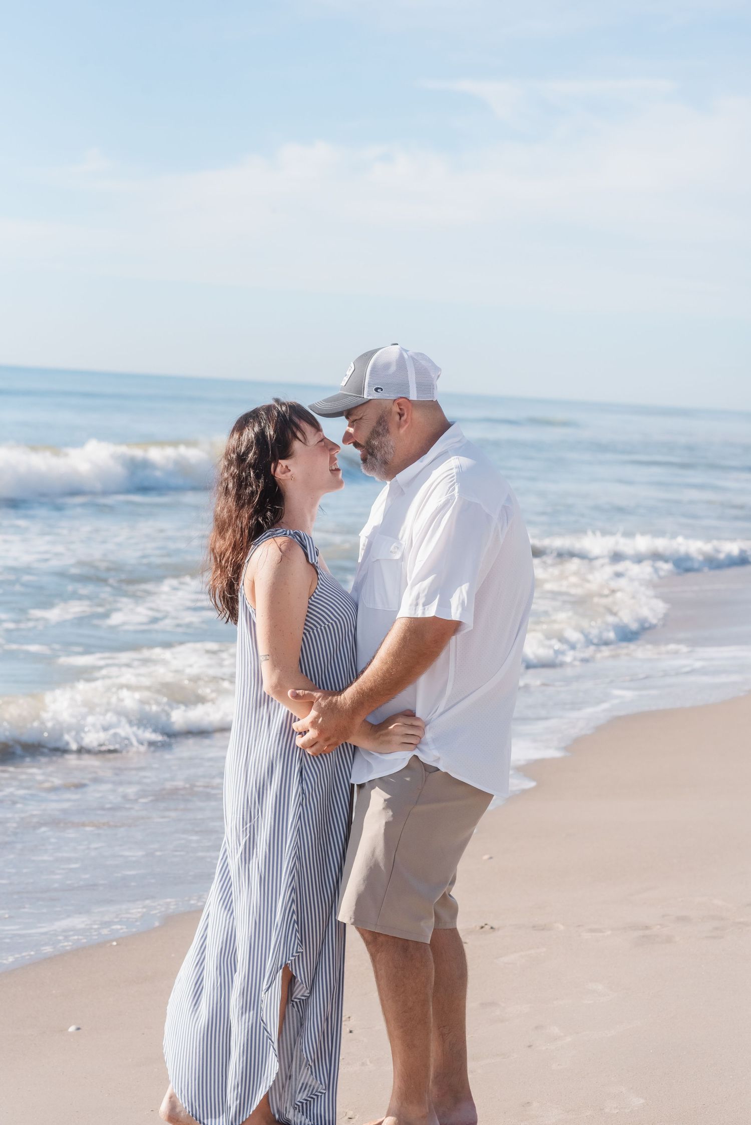 Couple holding hands and gazing at each other romantically on a sunny beach with ocean waves behind them.