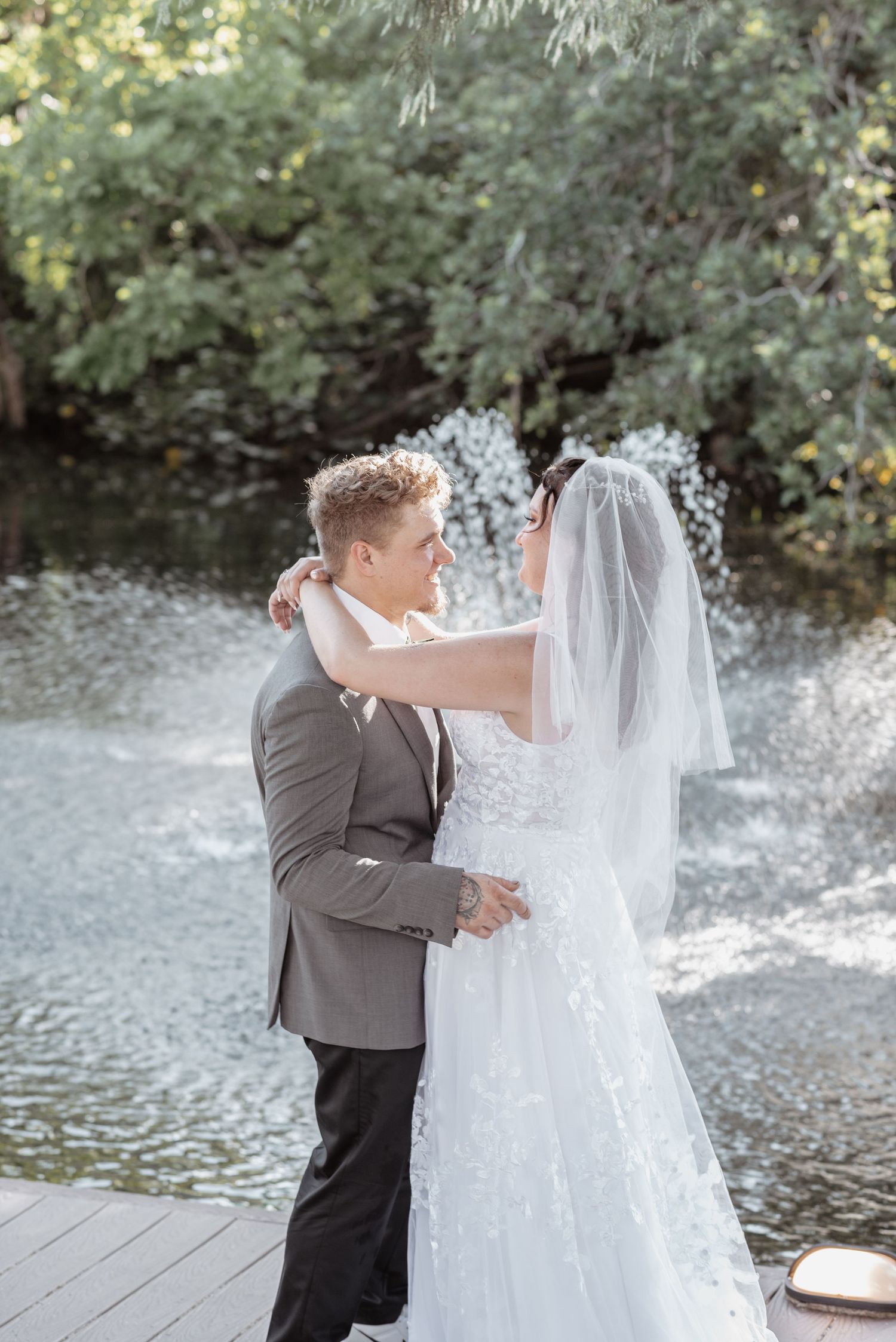 A bride and groom share a romantic embrace near a fountain and lush greenery on their wedding day.