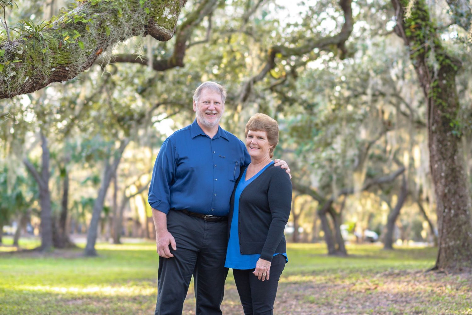 A smiling couple poses outdoors under moss-draped oak trees in a sunny park setting.