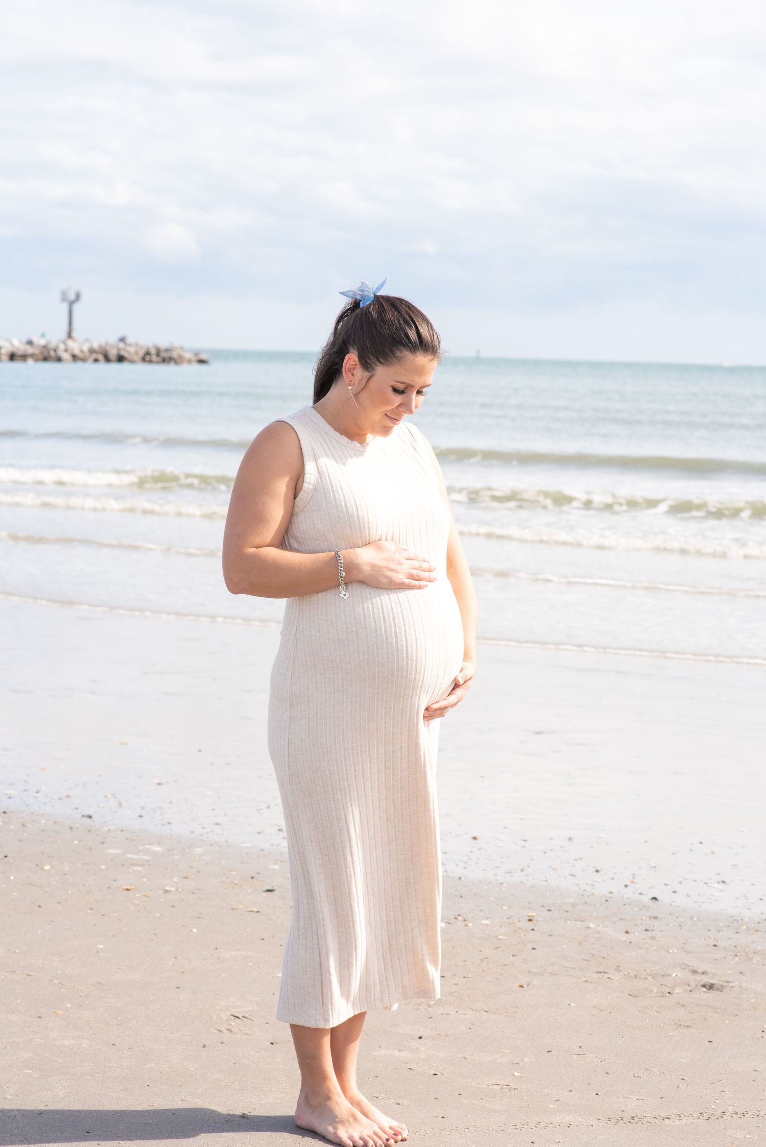 Pregnant woman in white ribbed dress cradling her baby bump on a sandy beach with ocean waves behind her.