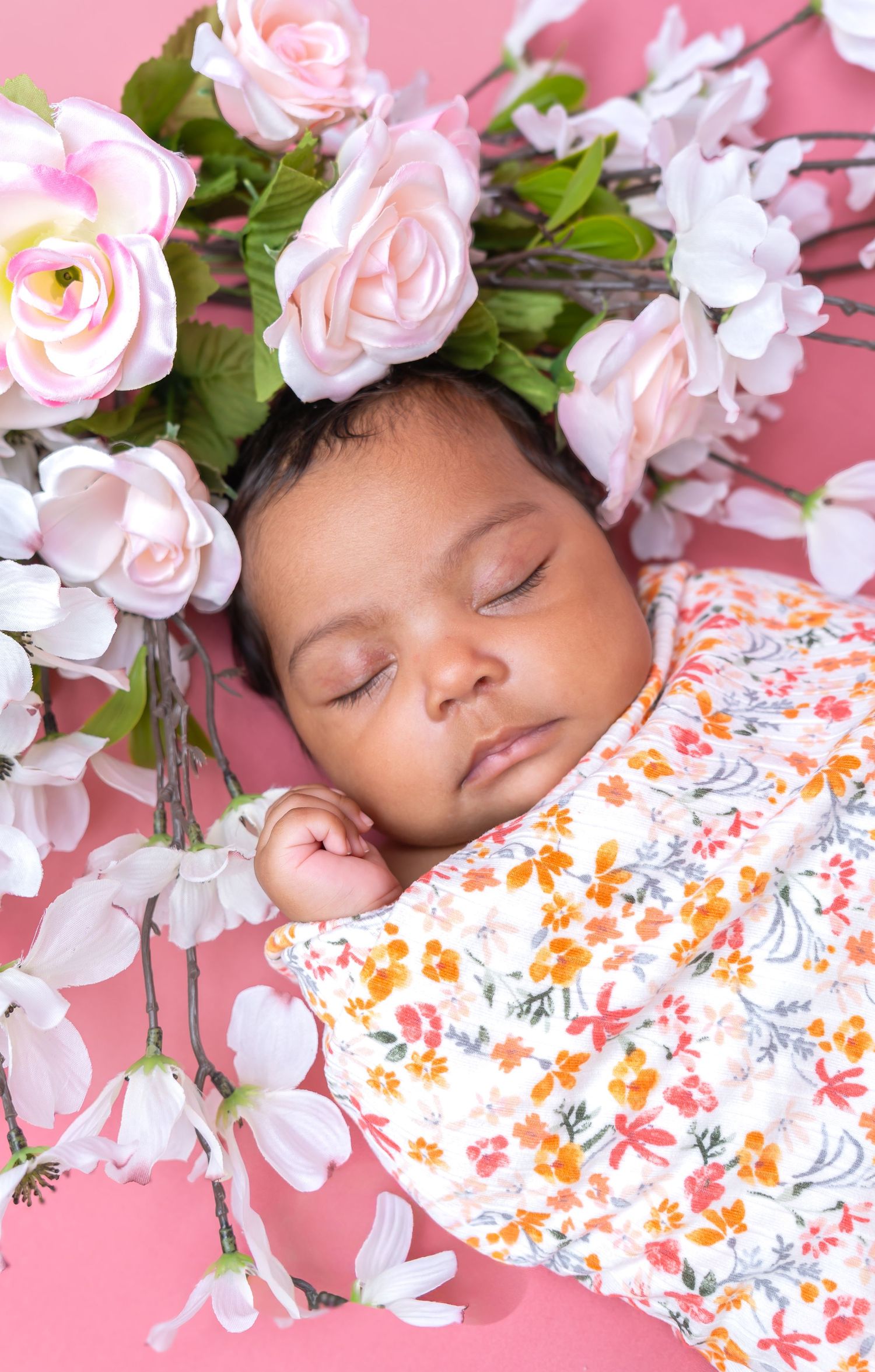 Sleeping newborn baby wrapped in floral swaddle surrounded by pink roses and white flowers on pink background.