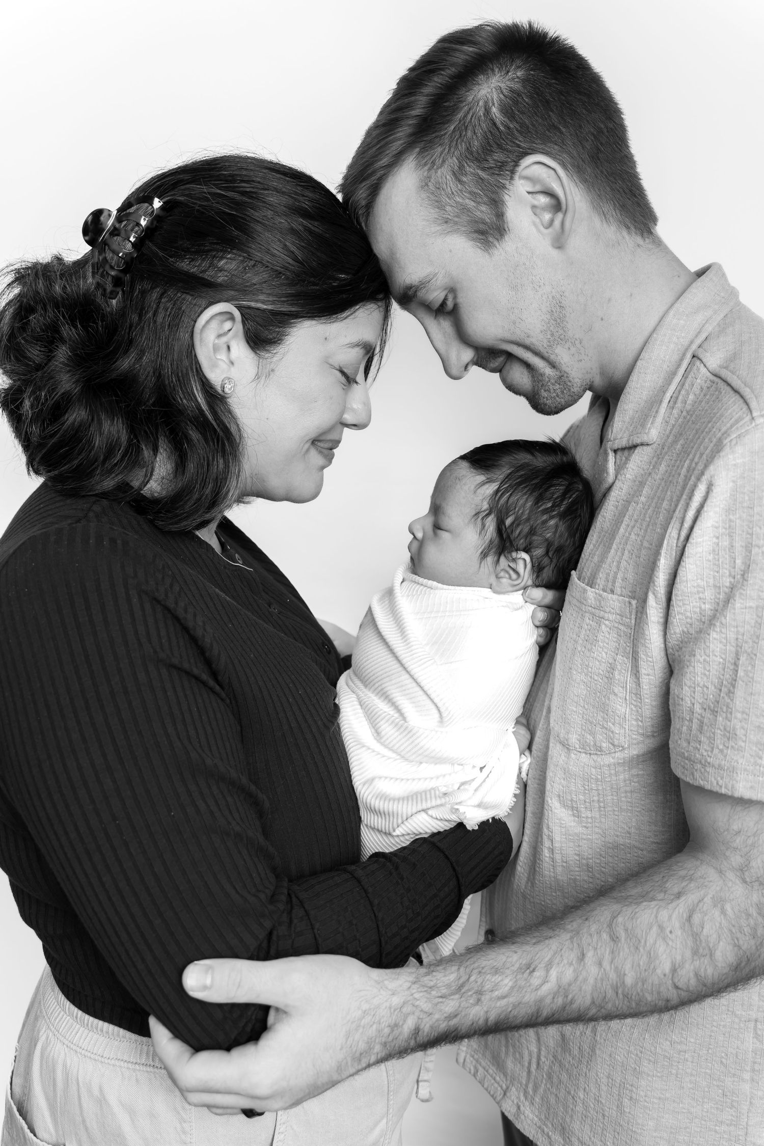 Black and white photo of new parents tenderly holding their newborn baby, foreheads touching in an intimate family portrait.
