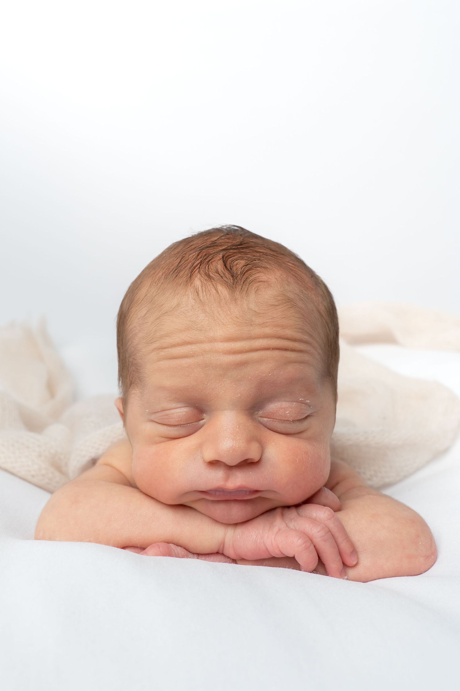 Close-up of a peaceful sleeping newborn baby resting chin on hands against white backdrop.