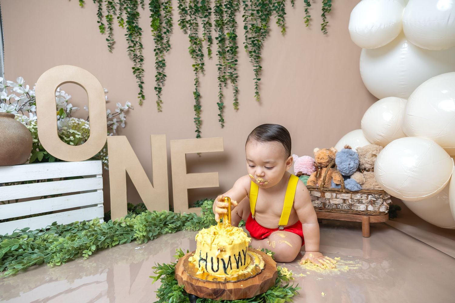 Baby boy in red swimsuit smashes yellow first birthday cake surrounded by greenery and ONE letter decor.