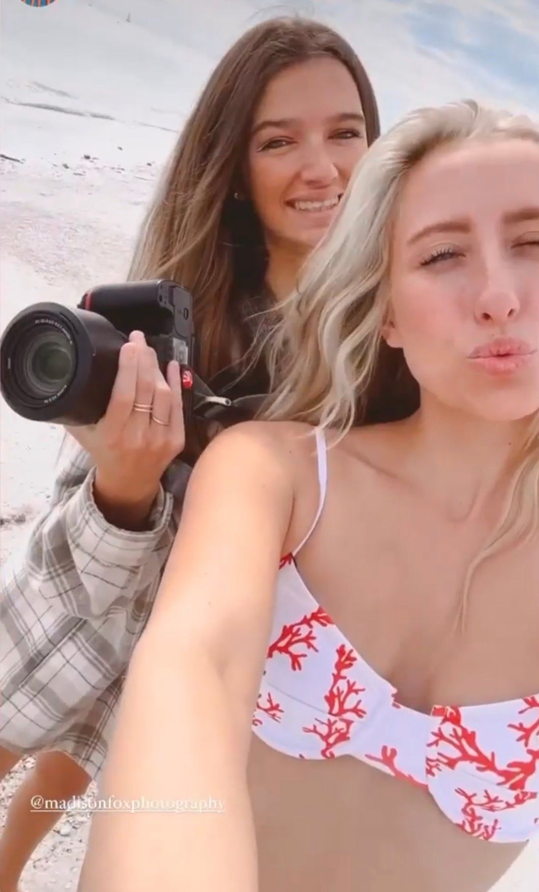 Female photographer holding camera poses with blonde woman in coral bikini top during beach photoshoot.