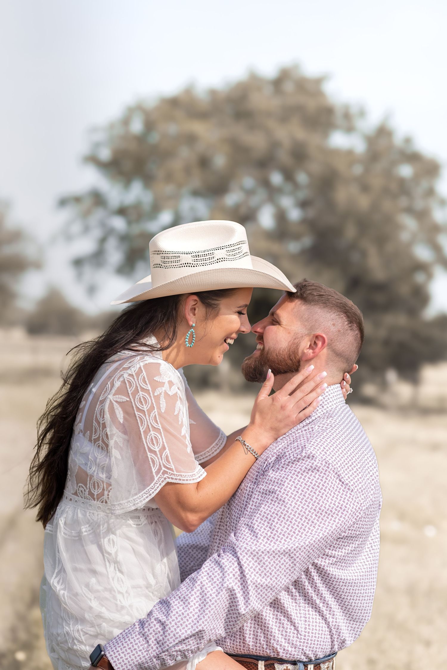 Couple sharing a romantic moment outdoors, woman in white lace dress and cowboy hat, man in plaid shirt.