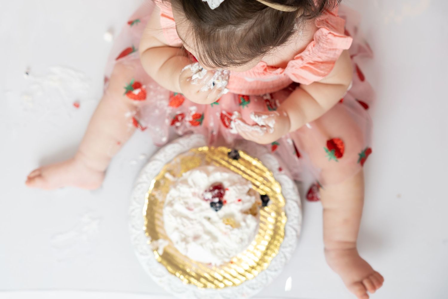 Baby girl in pink dress smashing her first birthday cake, viewed from above on white background.