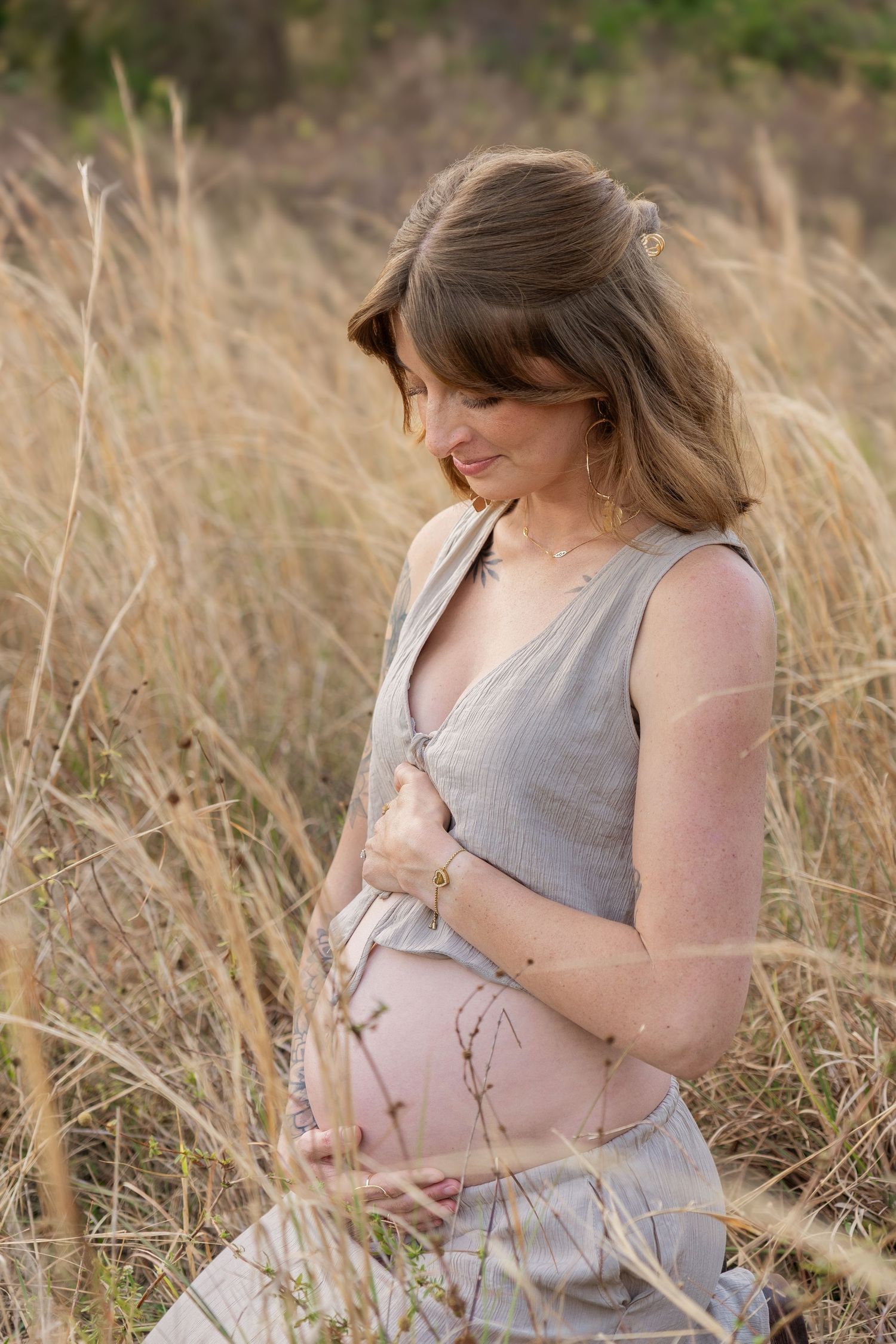 Pregnant woman in gray dress cradling her baby bump while standing in tall golden grass field outdoors.