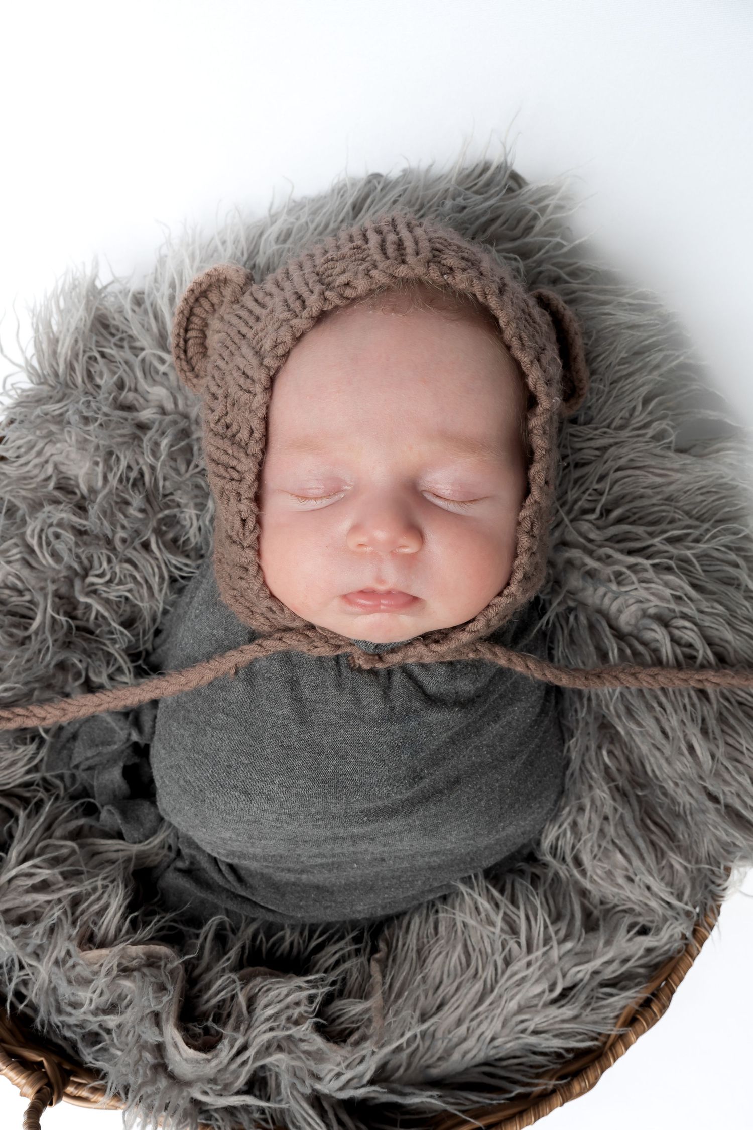 Sleeping newborn baby wrapped in gray swaddle wearing a brown knit bear bonnet hat, nestled in a fluffy gray basket.