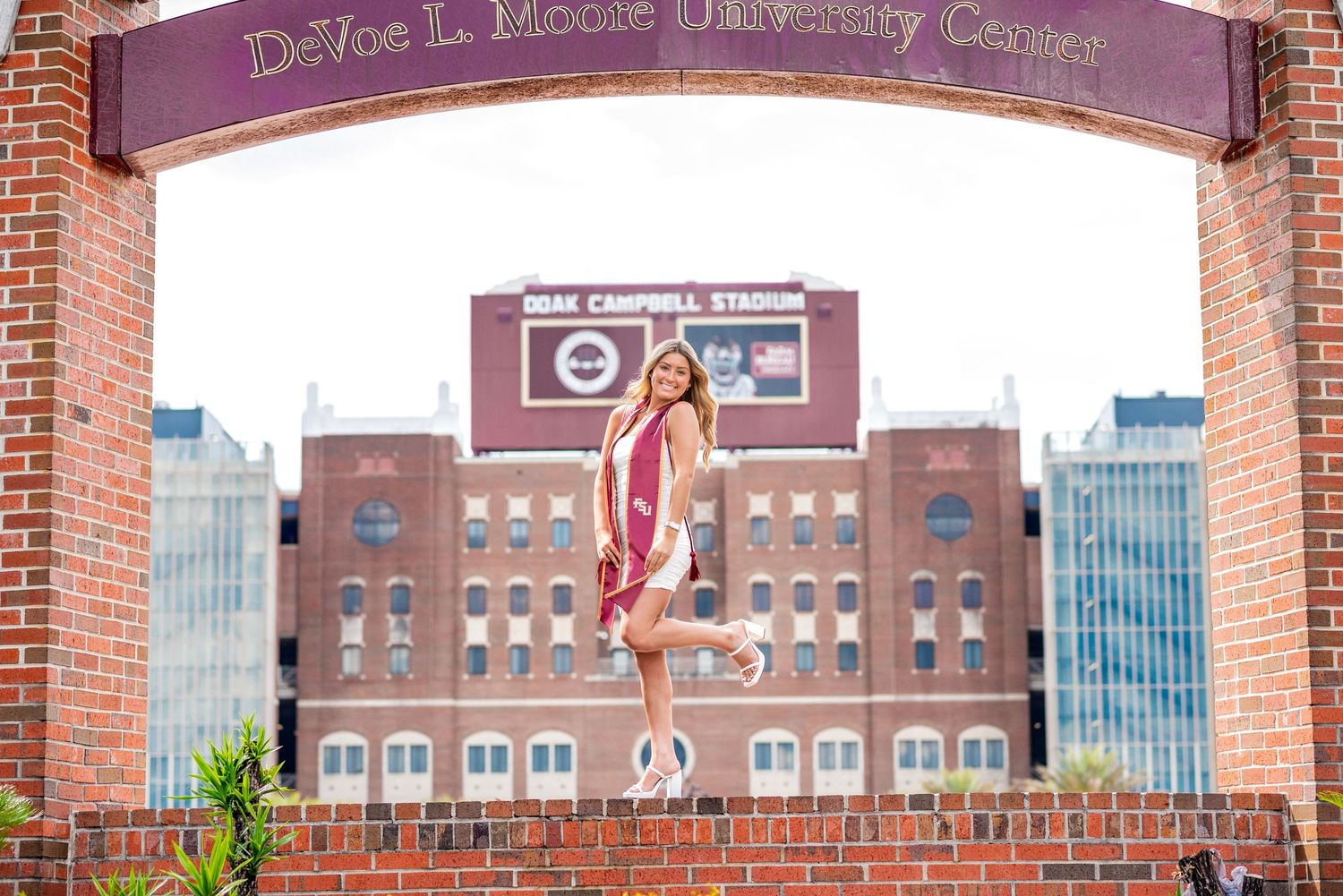 Graduate poses at DeVoe L. Moore University Center arch near Doak Campbell Stadium in maroon and gold regalia.