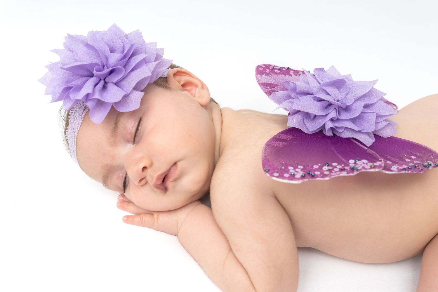 Sleeping newborn baby wearing purple flower headband and butterfly wings costume on white background.