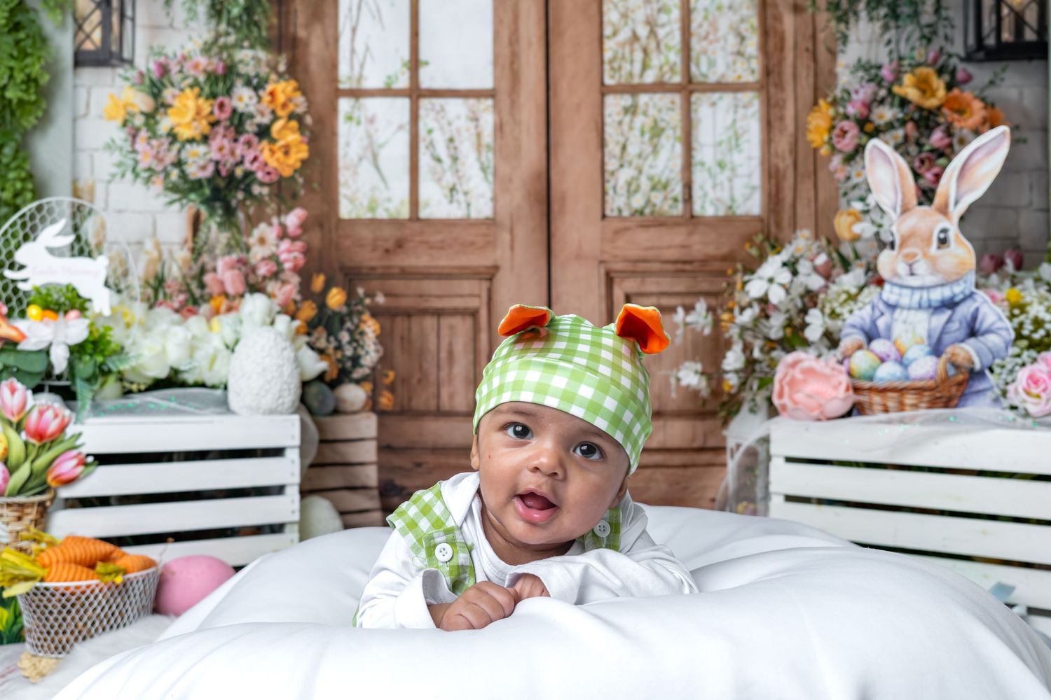 Smiling baby in green gingham hat poses on white blanket with Easter bunny and floral backdrop.