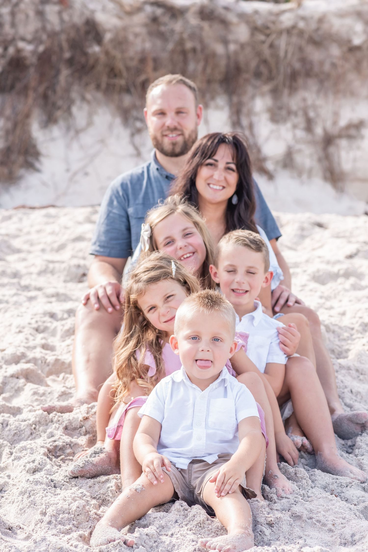 Happy family of six posing together on sandy beach, parents with four young children smiling outdoors.