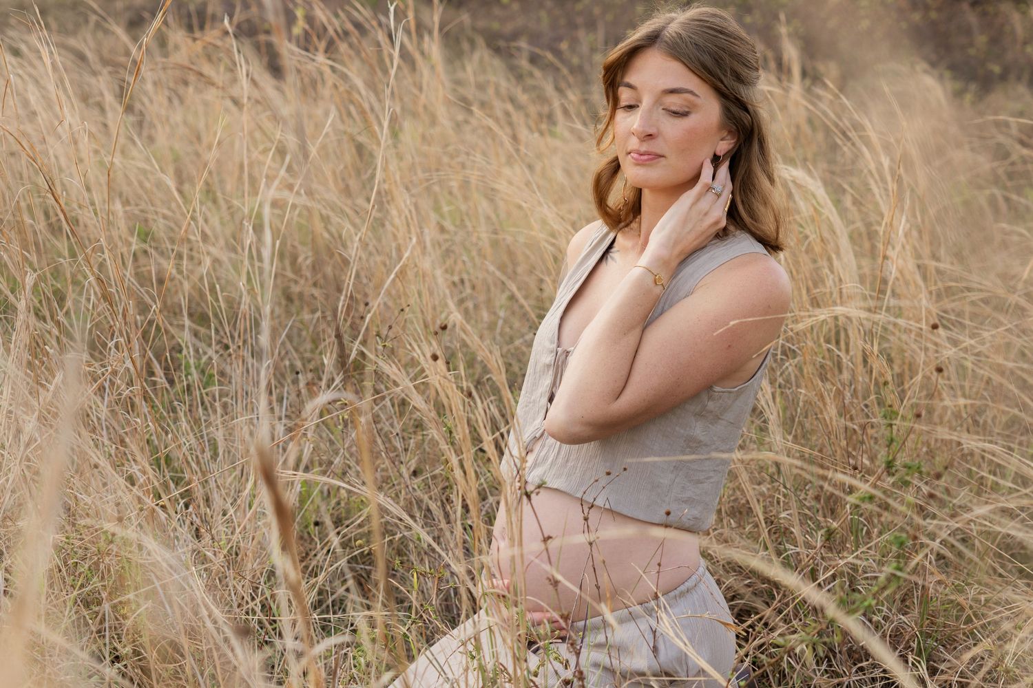 Pregnant woman in gray dress standing peacefully in golden tall grass field outdoors.