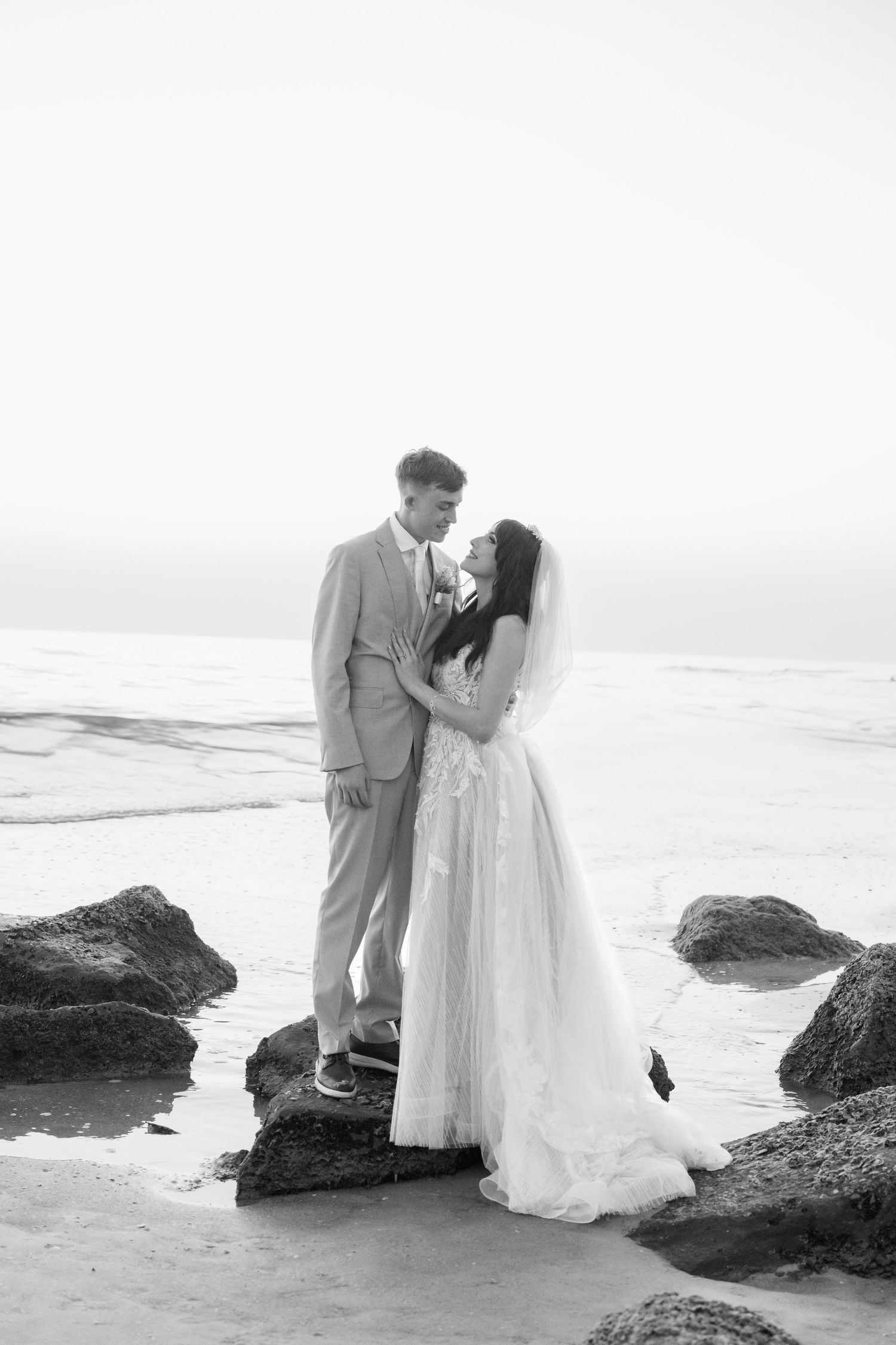 Black and white photo of a bride and groom standing on rocks at the beach, gazing at each other romantically.