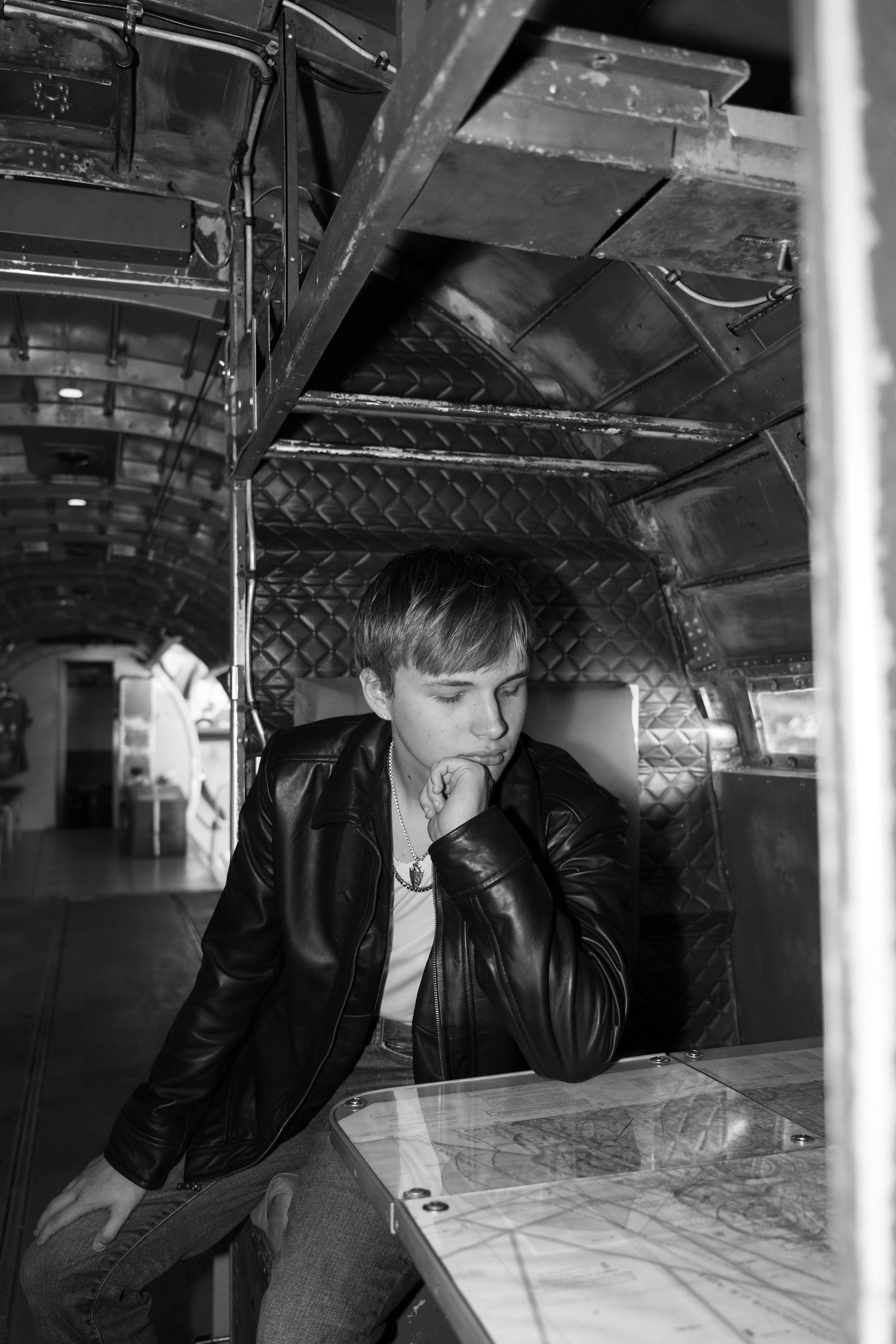 Young man in leather jacket sits pensively at a table inside a vintage aircraft interior, black and white photo.