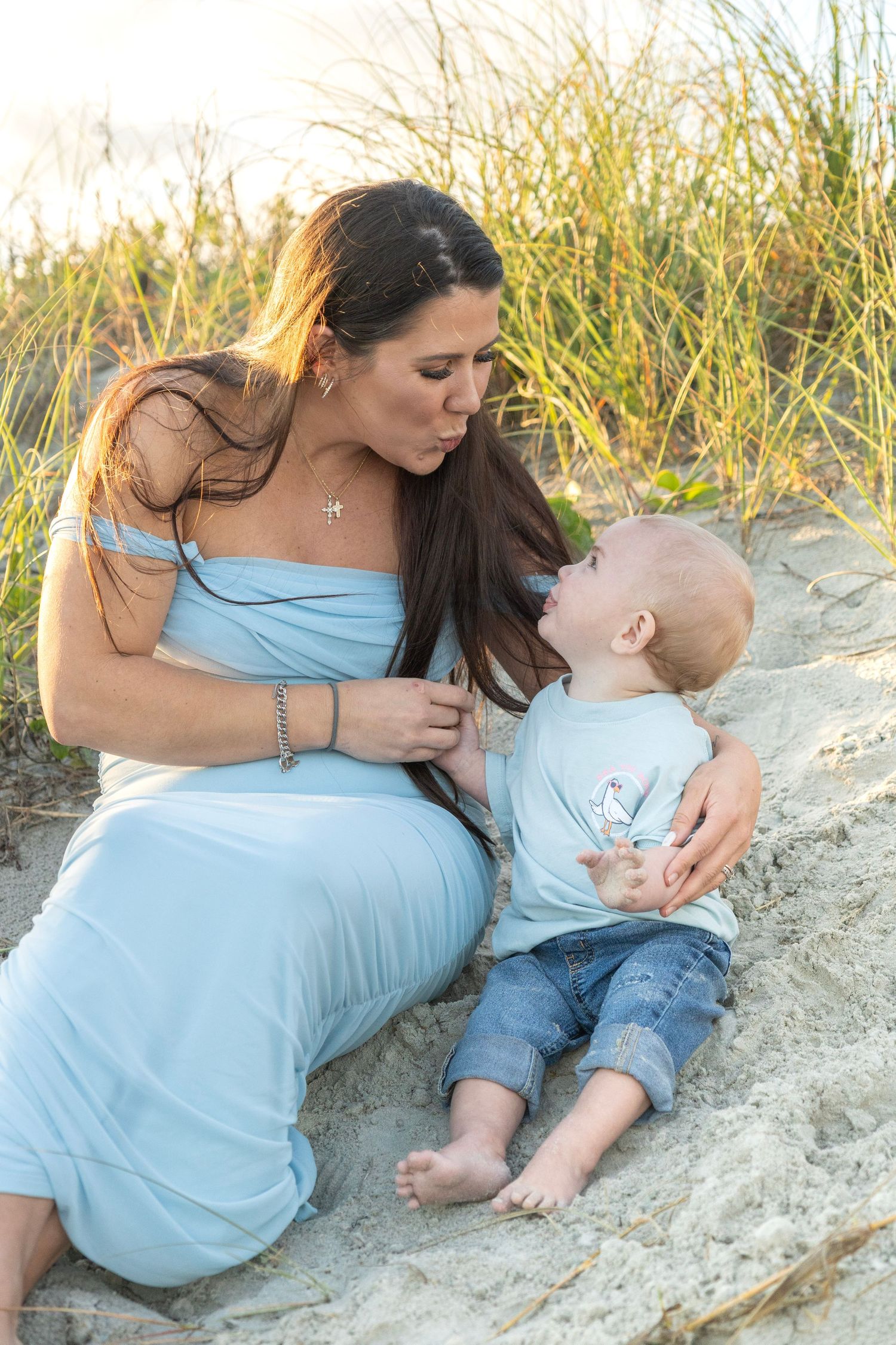 Pregnant woman in blue dress sitting on beach sand with toddler boy, sharing a tender moment outdoors.
