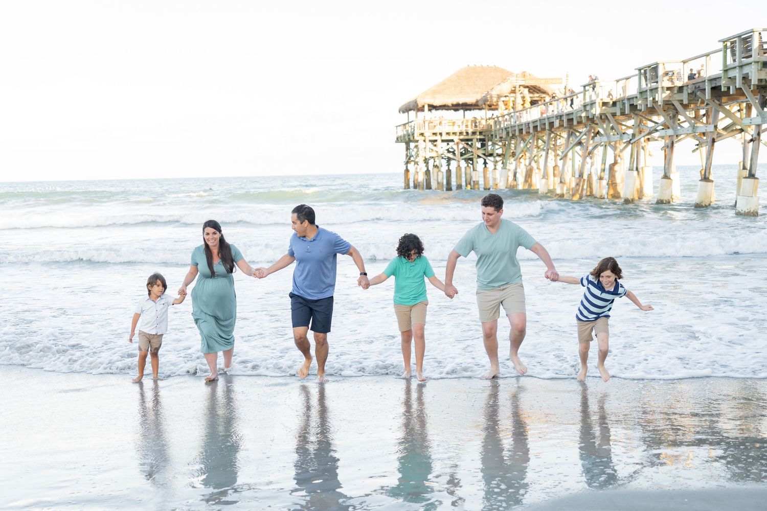 Happy family of five holding hands walking along ocean shoreline near a wooden beach pier.