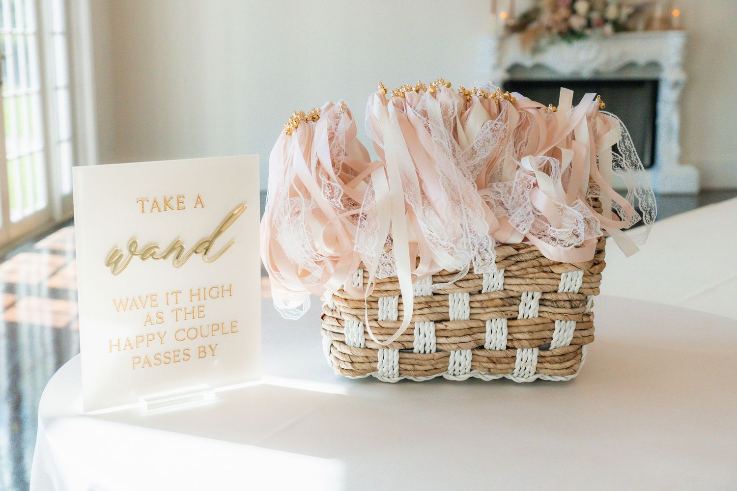 Wicker basket filled with blush ribbon wands next to a gold sign reading Take a Wand at a wedding reception.