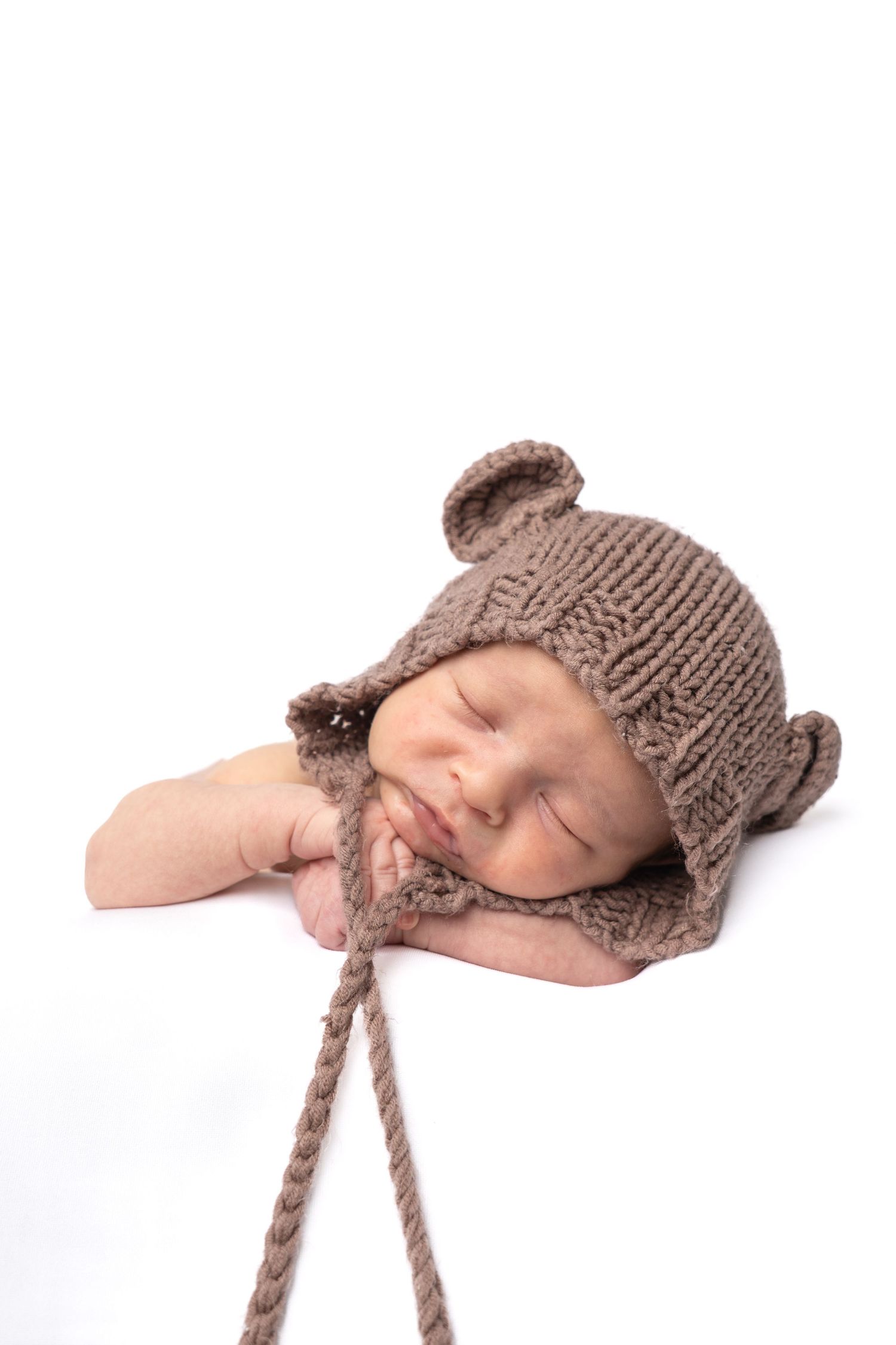 Sleeping newborn baby wearing a brown knitted bear ear hat on a white background.