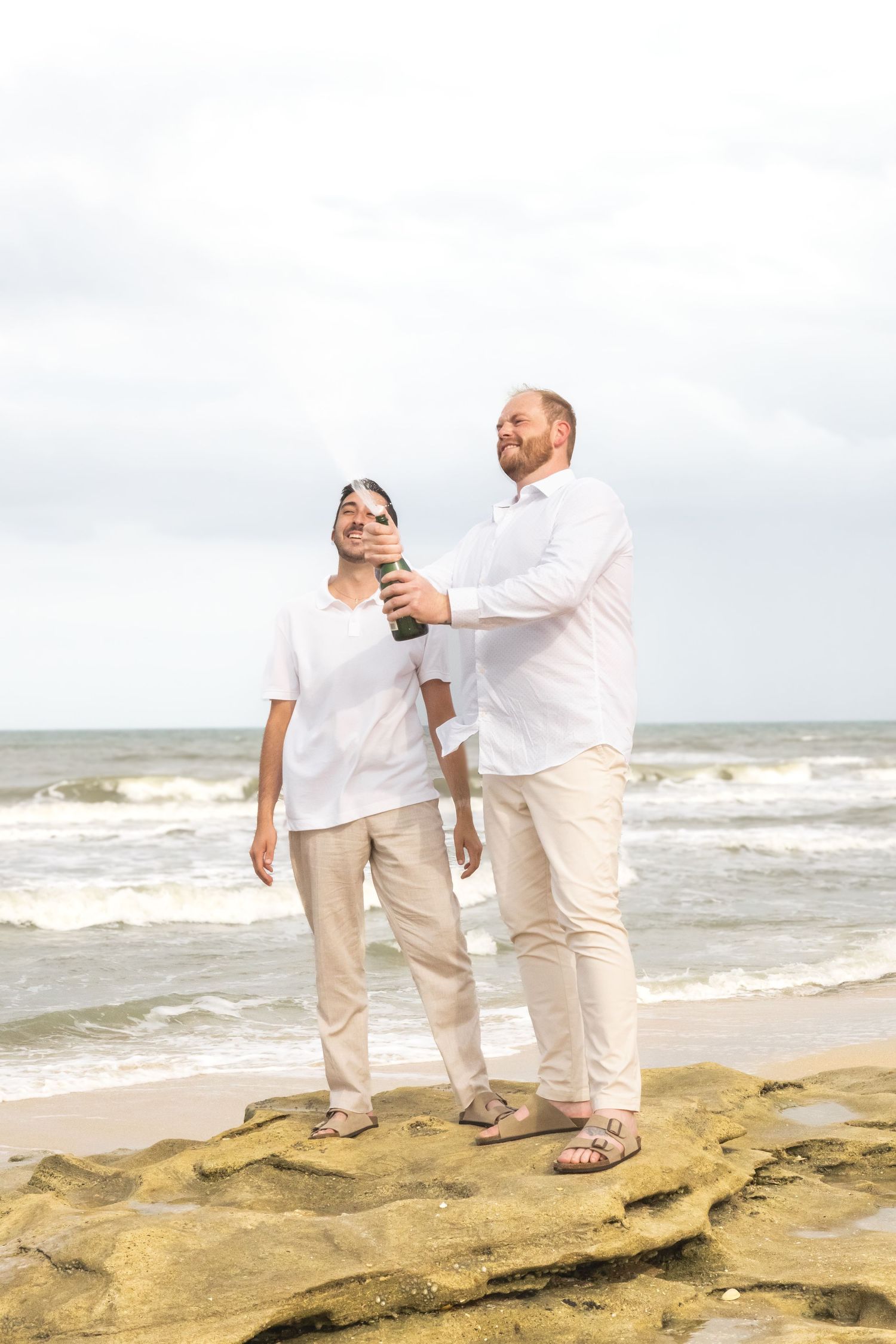 Two men in white shirts celebrate on a rocky beach, sharing a champagne bottle with ocean waves behind them.