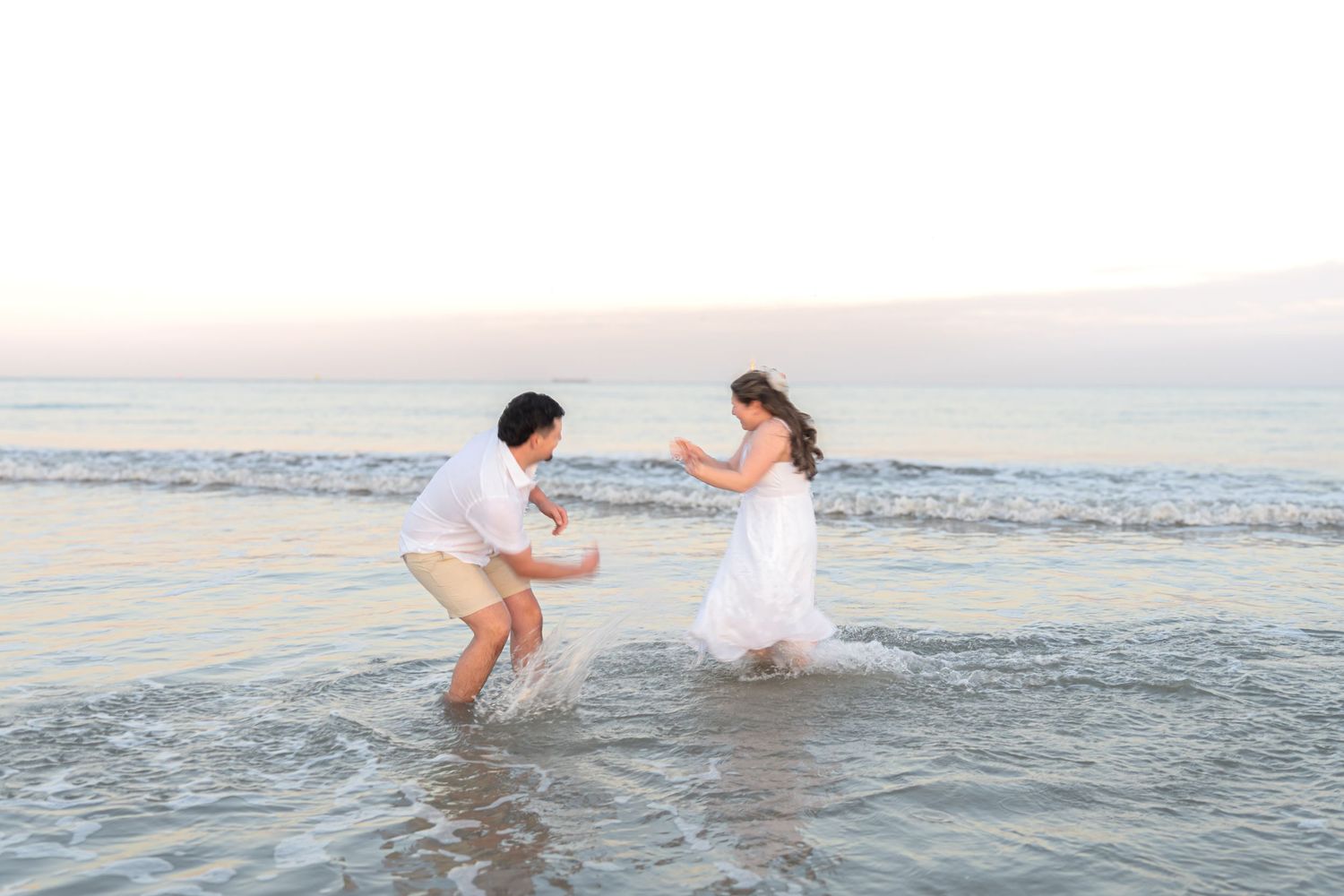 Bride and groom splashing in ocean waves at sunset during a romantic beach wedding photo session.