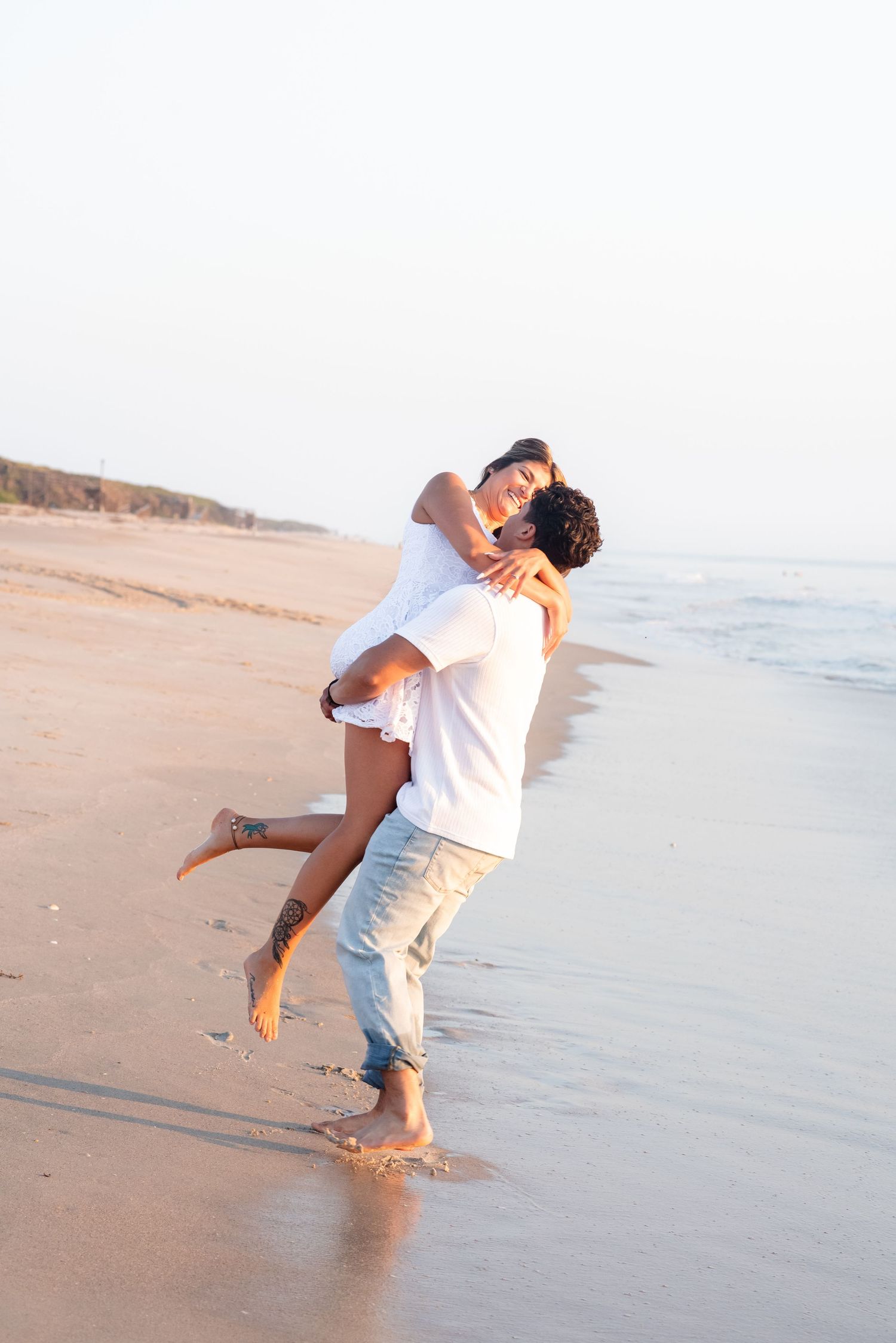 Romantic couple embracing on a sandy beach at sunset, man lifting woman in white dress near ocean waves.