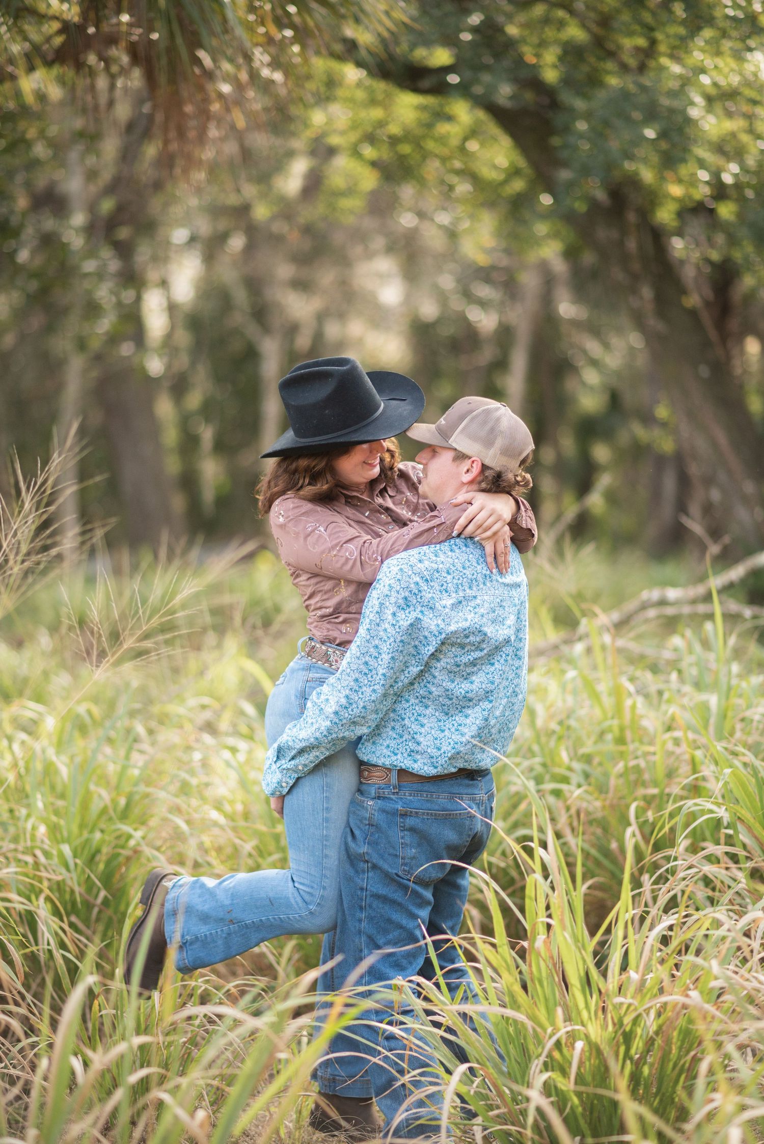 Couple in cowboy hats embracing in tall grass field, man lifting woman in romantic outdoor country setting.