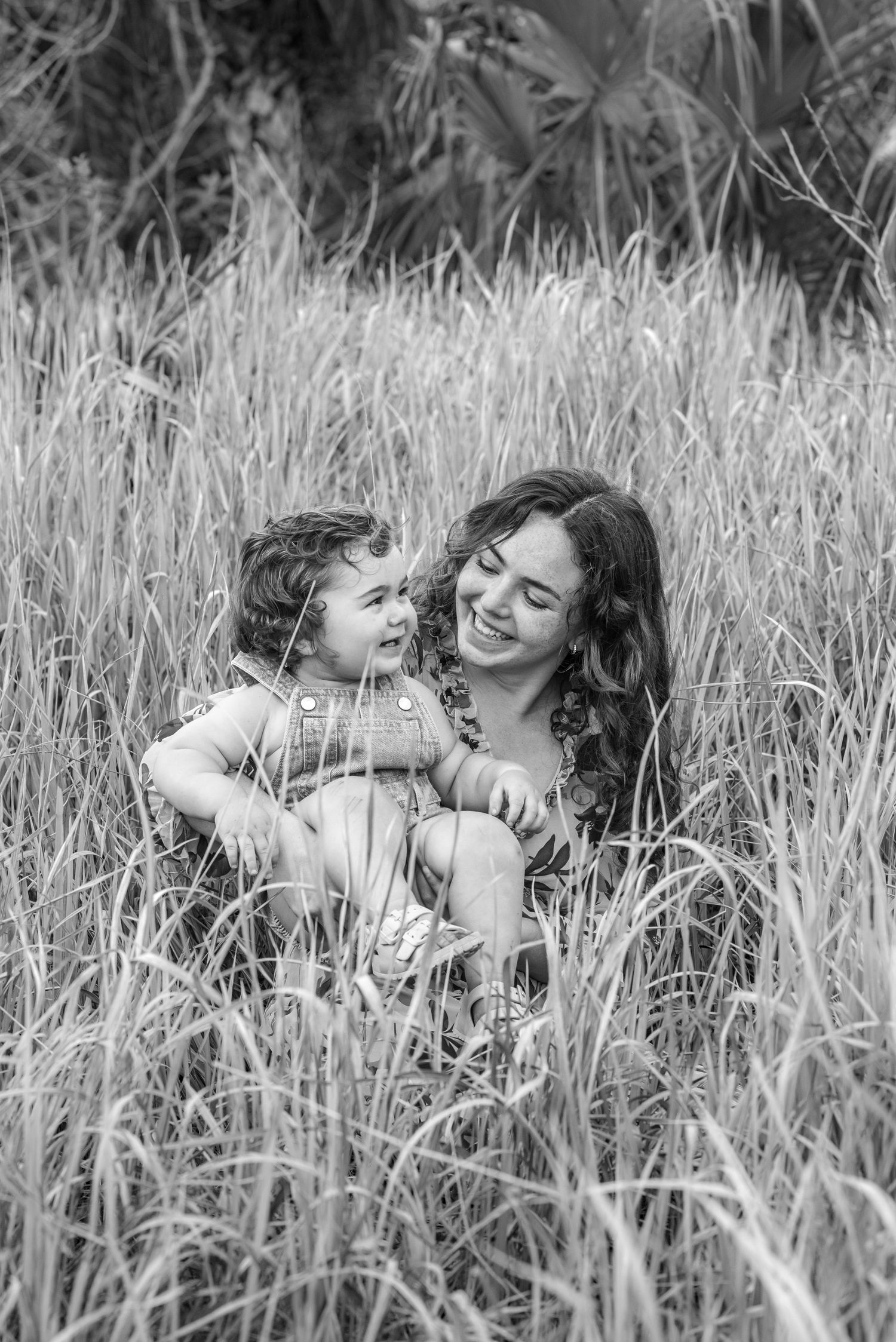 Black and white photo of a smiling mother sitting in tall grass holding her happy toddler child outdoors.