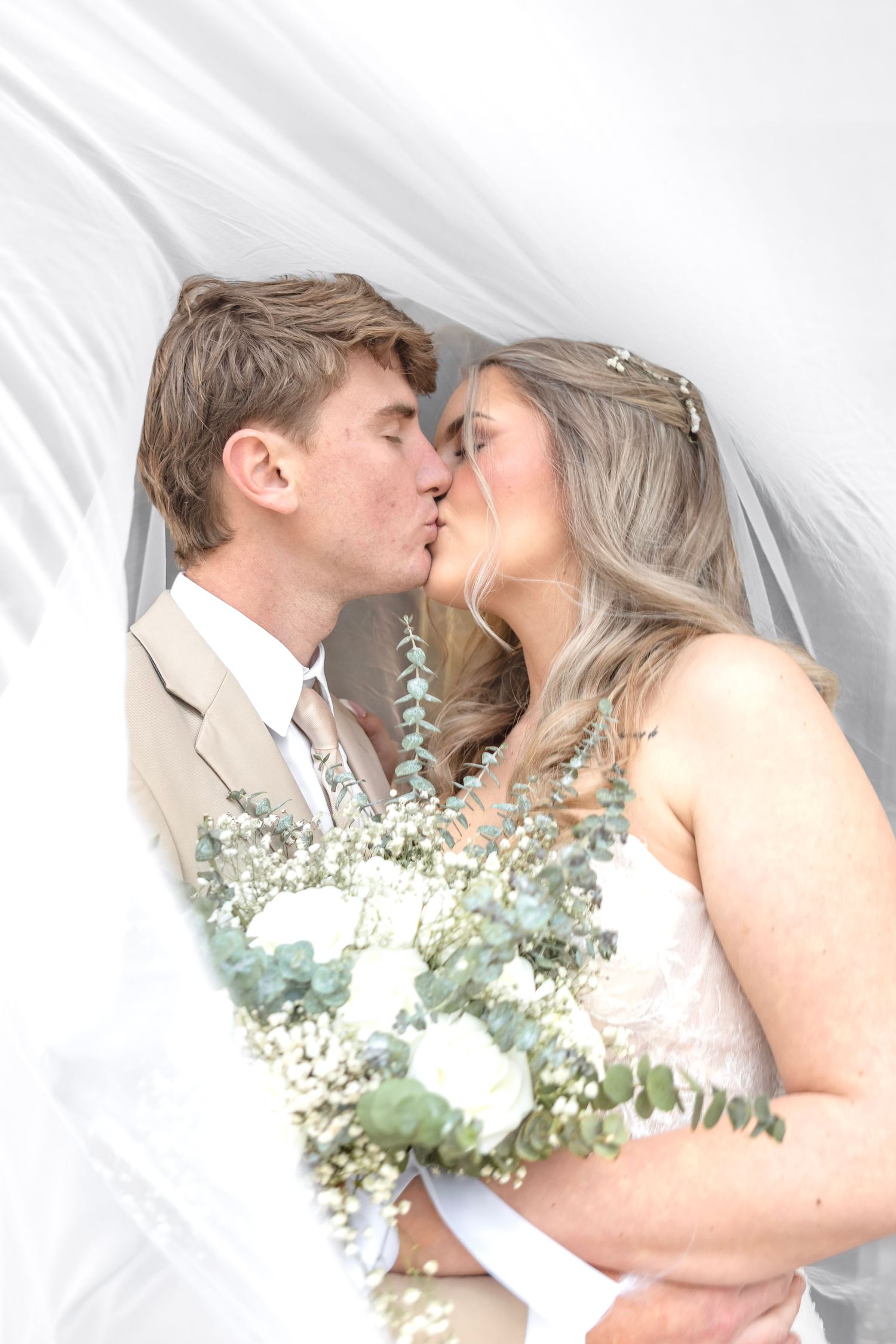 Bride and groom sharing a romantic kiss under a white veil, bride holding a bouquet of white roses and eucalyptus.
