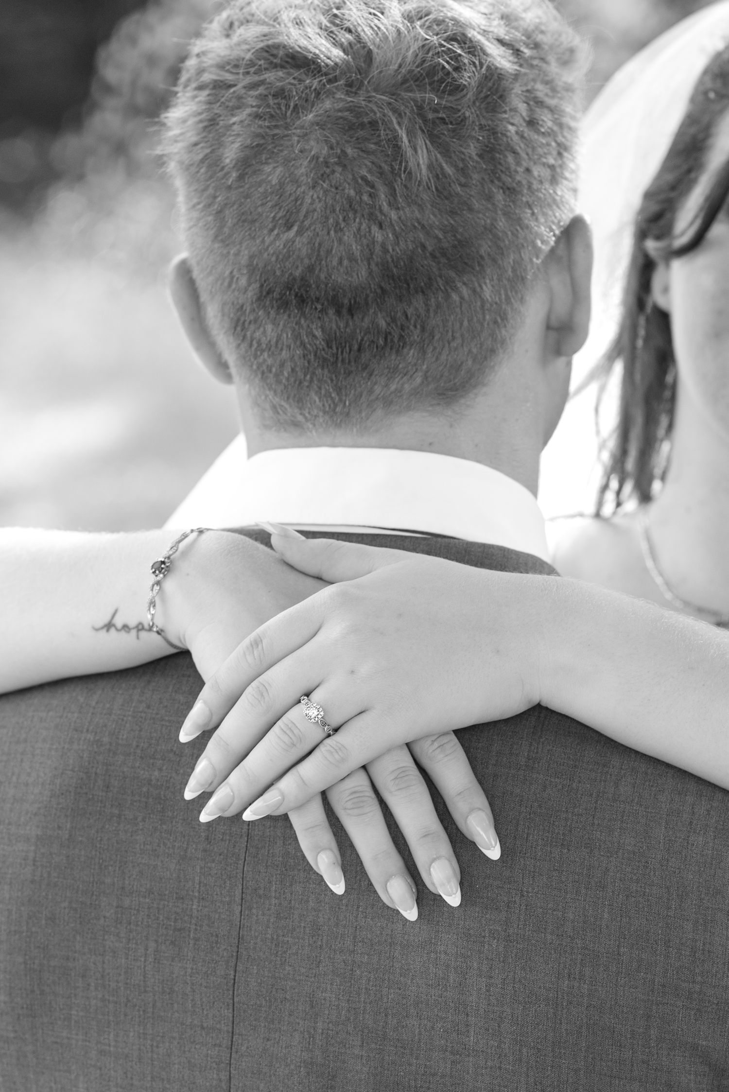 Black and white photo of bride resting hand with engagement ring on groom's shoulder at wedding.