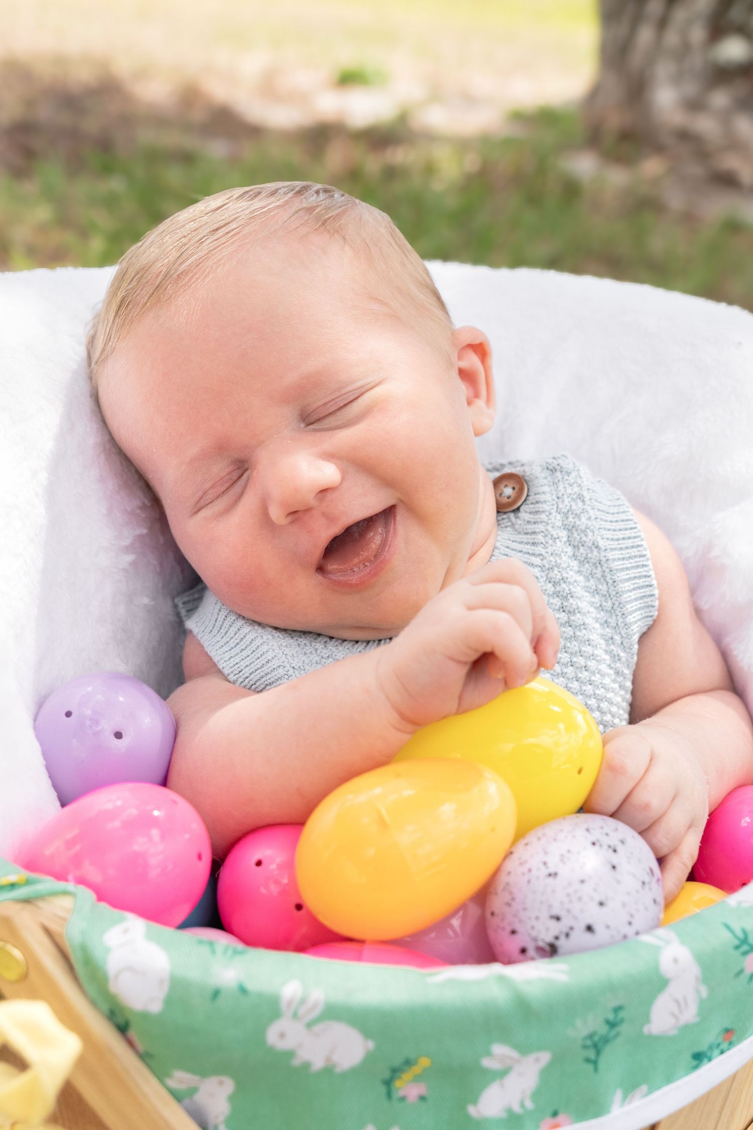 Laughing baby sitting in Easter basket surrounded by colorful plastic Easter eggs outdoors.